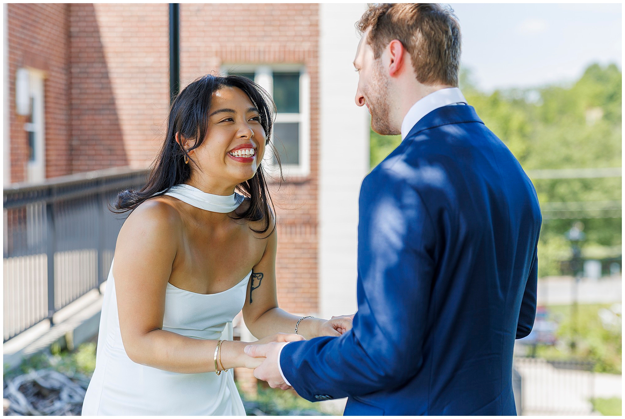A woman in a white dress smiles joyfully while holding hands with a man in a blue suit outdoors, celebrating their Beacon City Hall elopement against a backdrop of a brick building and lush greenery.