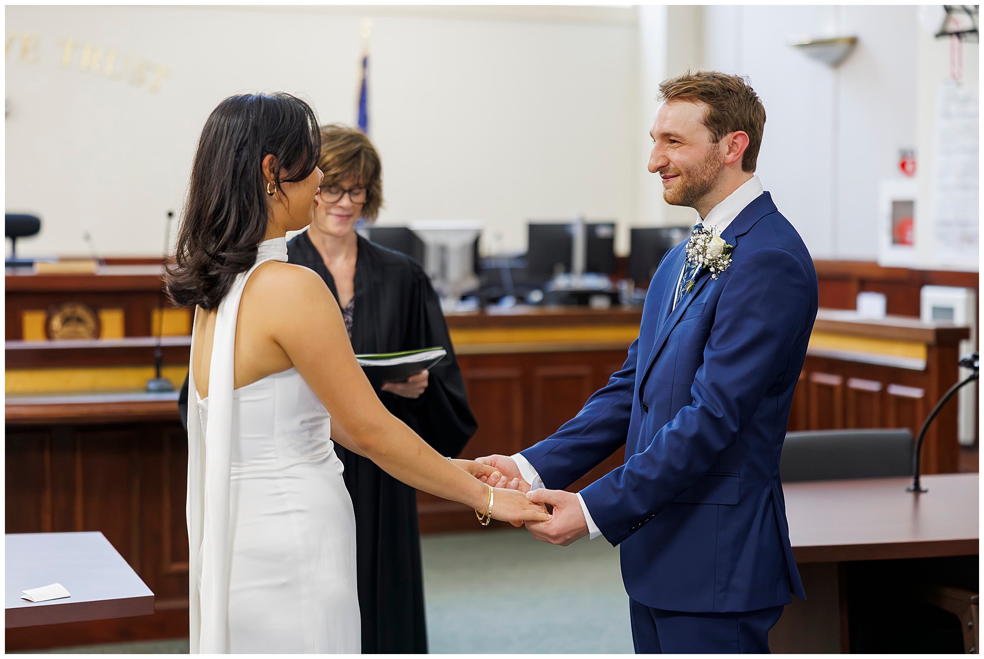 A couple holds hands and smiles at each other during their Beacon City Hall elopement ceremony, with an officiant standing behind them, holding a book.