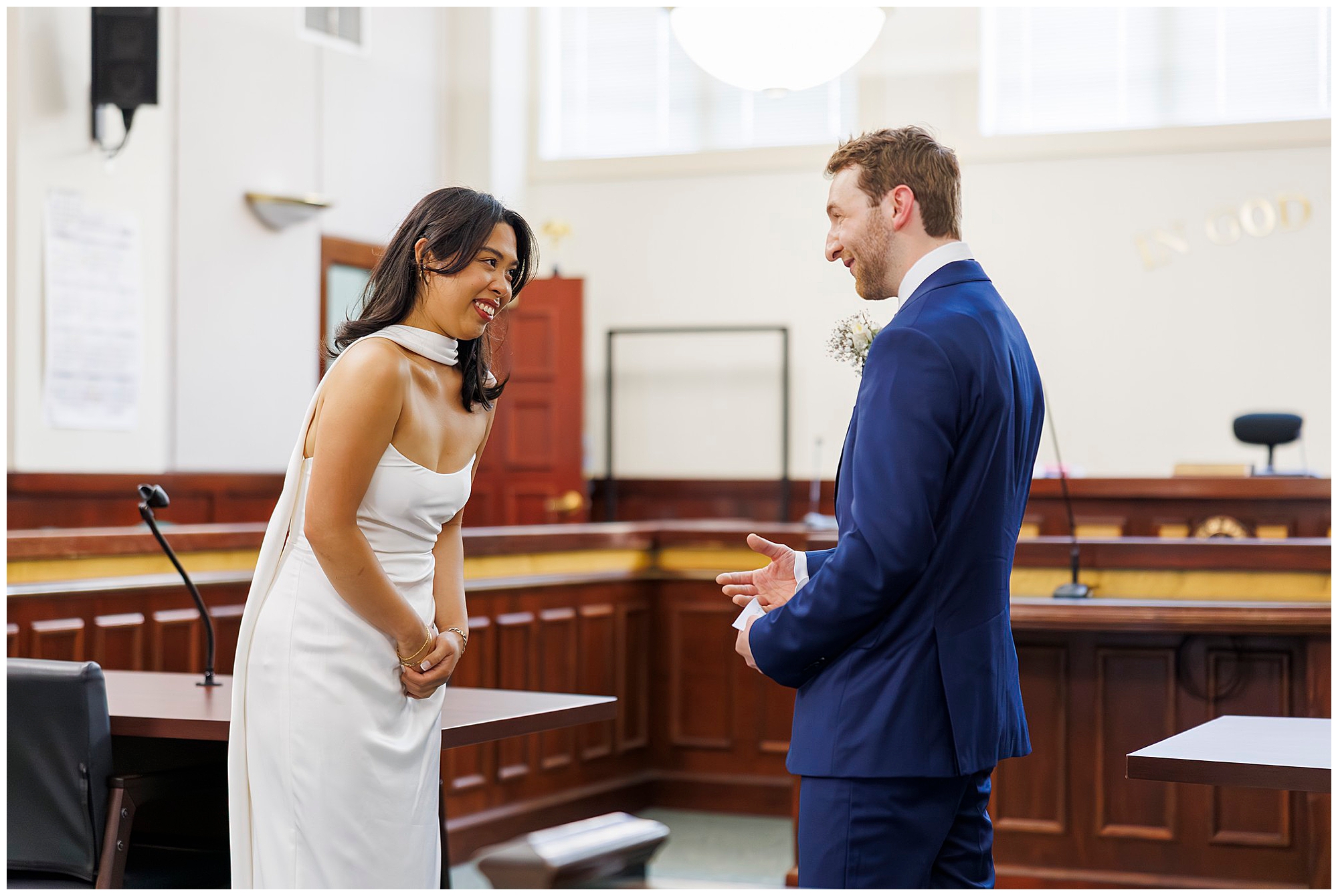 A woman in a white dress and a man in a blue suit smile and talk to each other inside Beacon City Hall, standing near desks with a judge’s bench visible—capturing the joy of their intimate elopement.