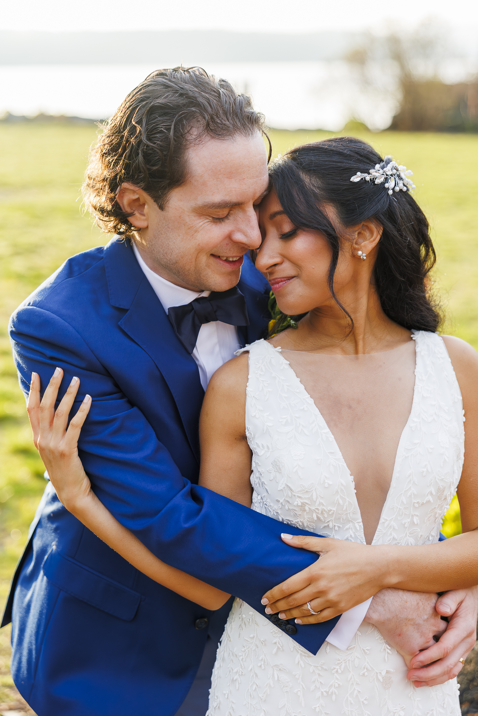 A newlywed couple embraces outdoors at their Harvest on Hudson micro wedding. The groom in a blue suit hugs the bride in a white, sleeveless dress with a deep neckline and hairpiece. Smiling with eyes closed, they share a tender moment.