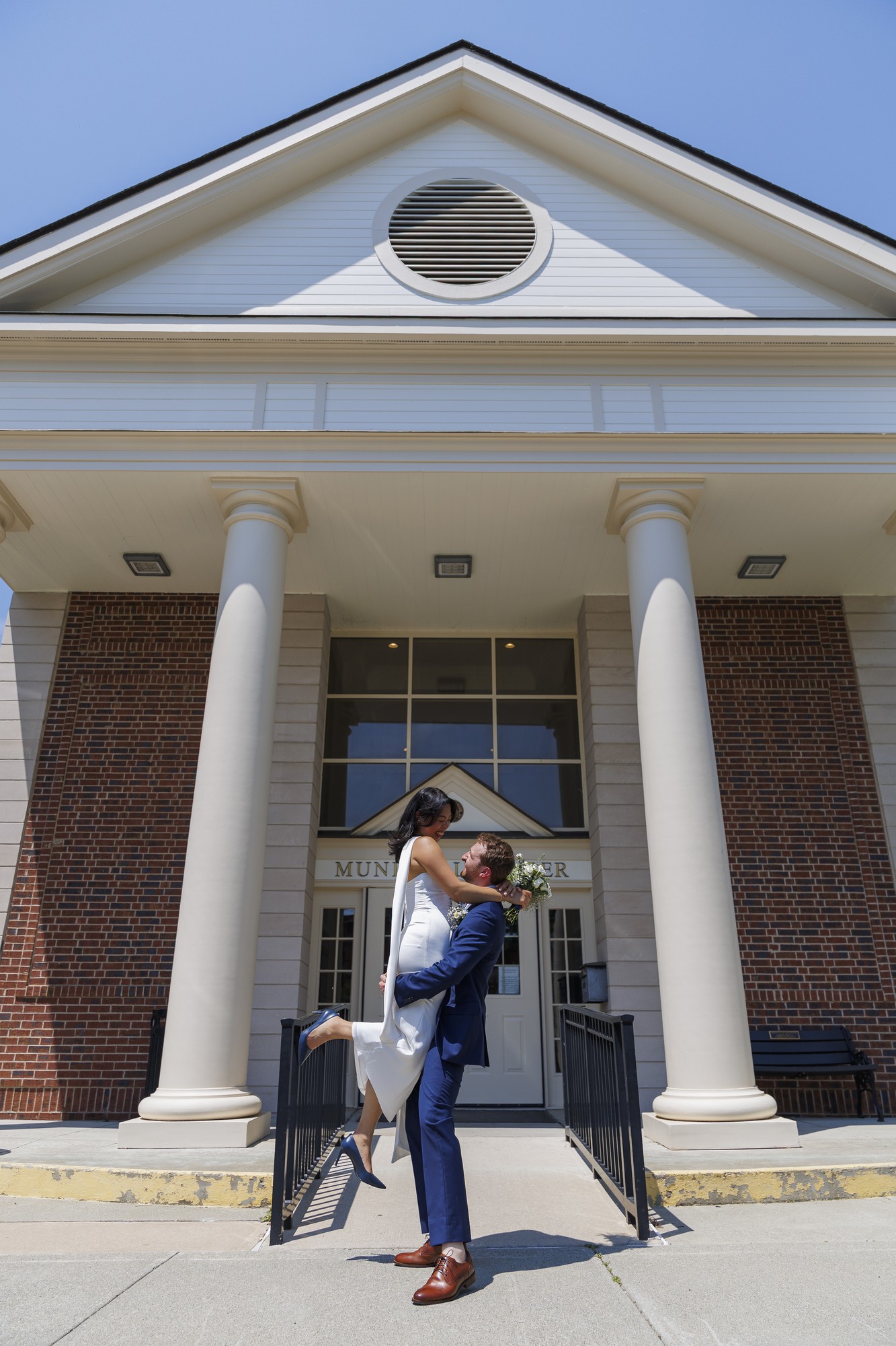 A groom in a blue suit lifts his bride, dressed in white, outside a building with tall columns and a brick facade on a sunny day. The couple is smiling and looking at each other lovingly.