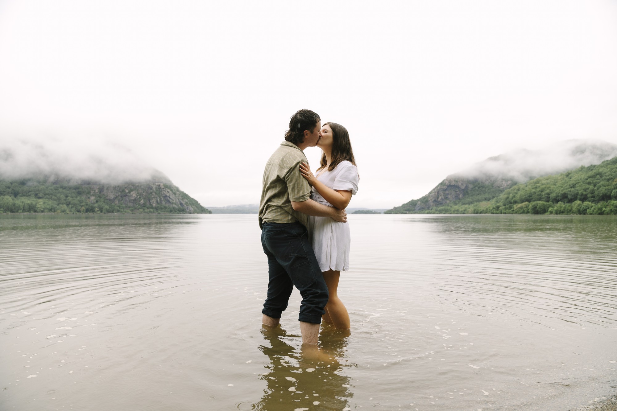 A couple stands in shallow water, kissing and embracing during their Little Stony Point engagement photo shoot, surrounded by misty mountains and a calm lake on a cloudy day.