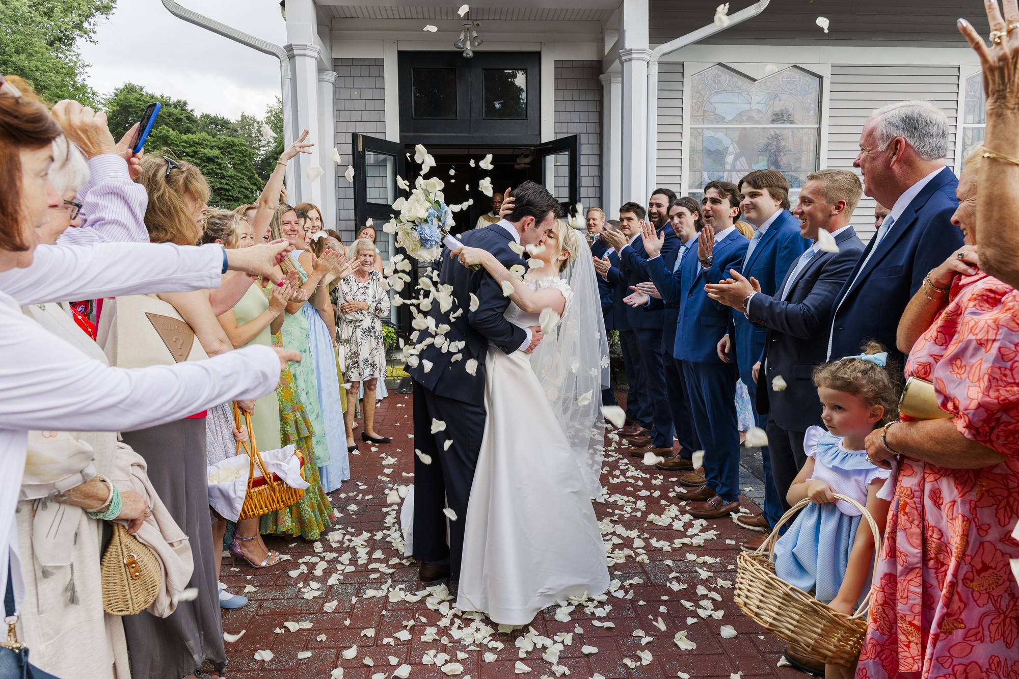 A bride and groom kiss outside the Church of the Magdalene as guests shower them with white flower petals. People line both sides, cheering and smiling, while a young girl in a blue dress holds a basket in the foreground.