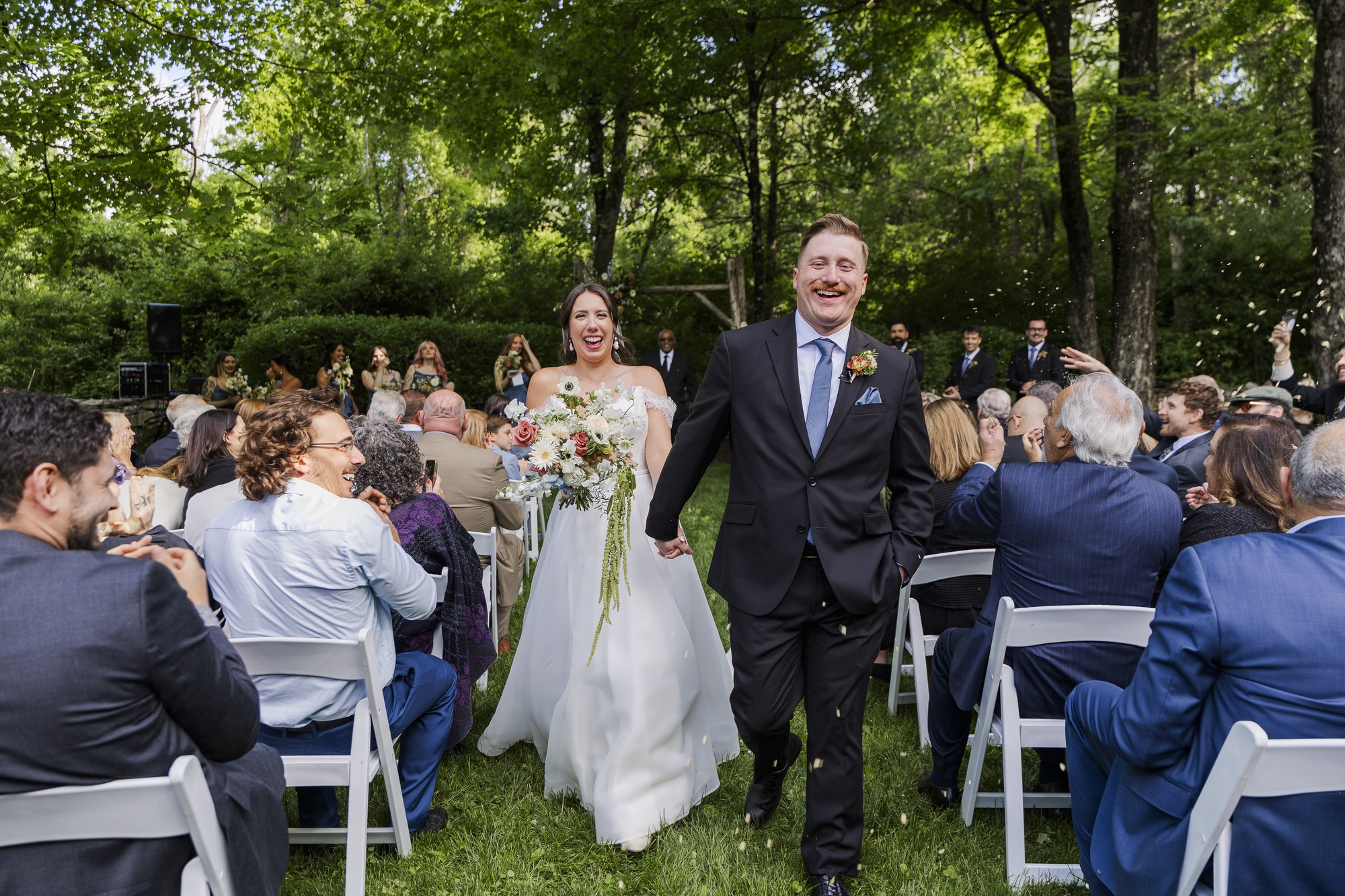 A bride and groom walk joyfully down an outdoor aisle at their Preston Barn wedding, hand in hand, surrounded by seated guests on white chairs. Lush green trees and the wedding party are visible in the background.