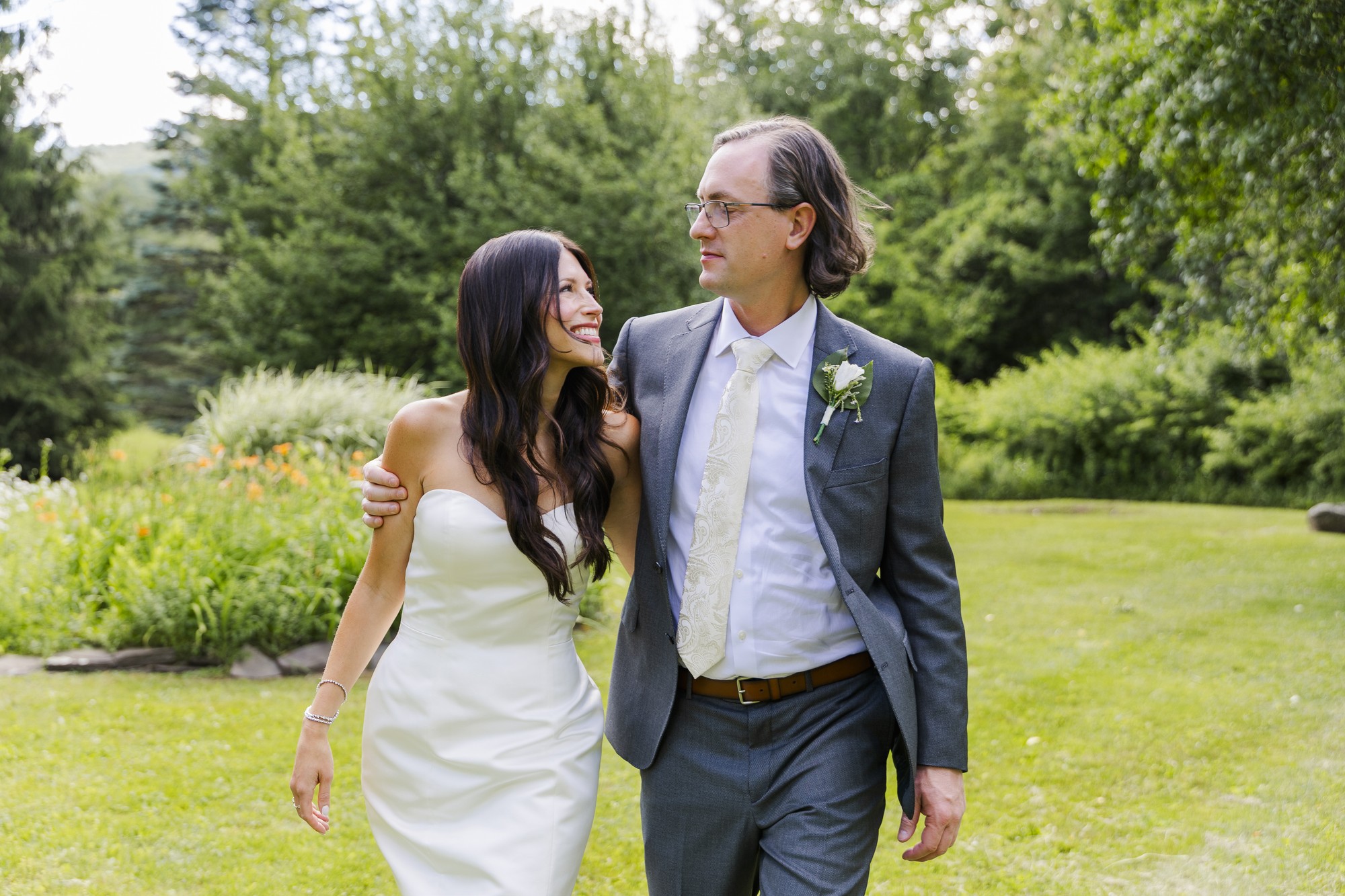 A bride in a white dress and a groom in a gray suit walk arm in arm outdoors, smiling at each other in a lush green garden setting.