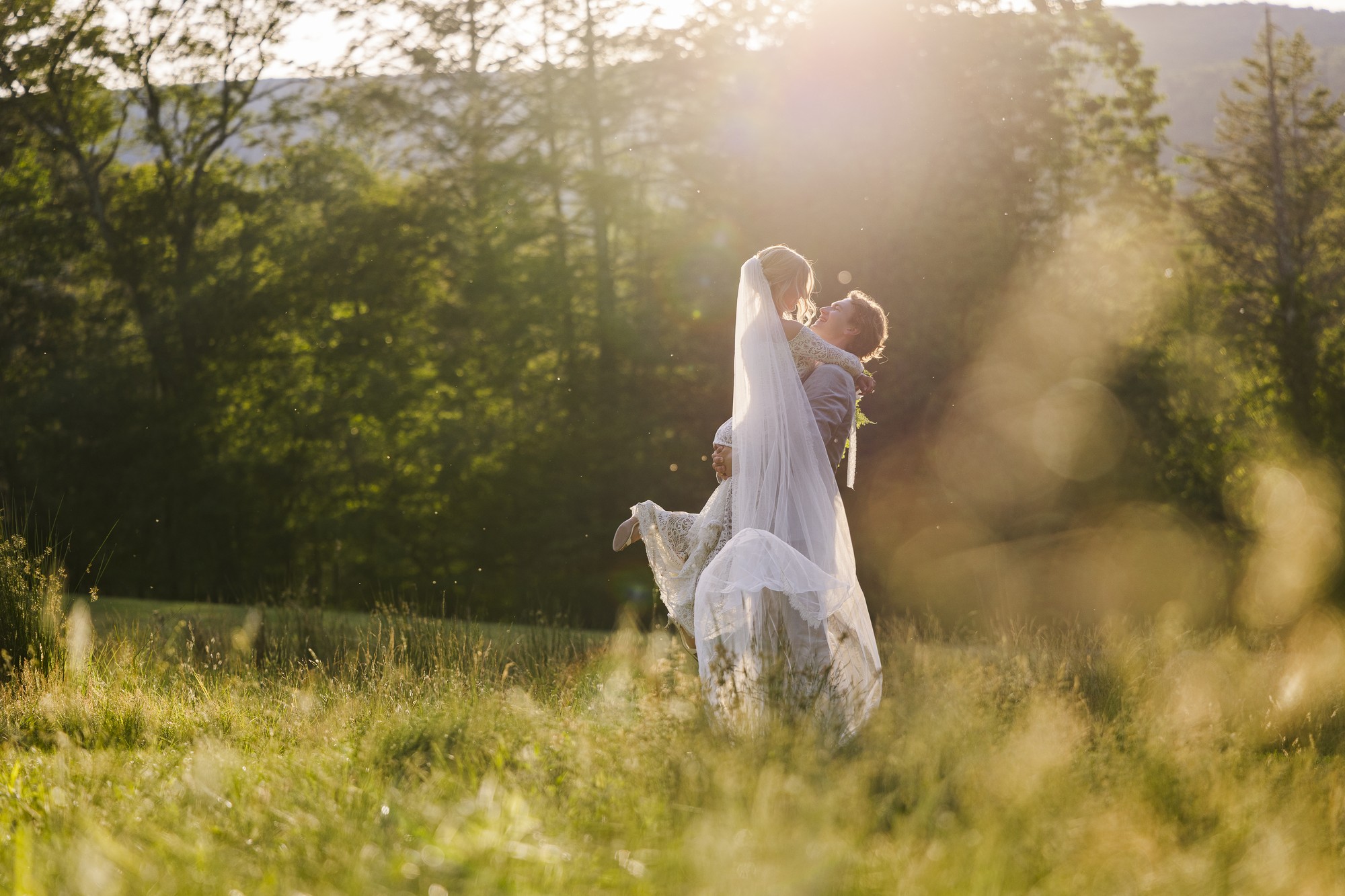 A bride and groom stand in a sunlit field, with the groom lifting the bride playfully. Tall grass surrounds them, and trees and hills are visible in the bright, golden background.
