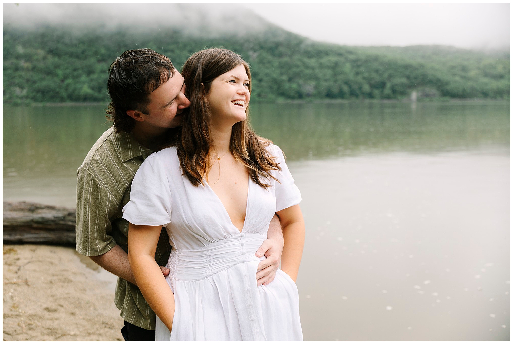 A man hugs a smiling woman from behind as they stand on a lakeshore during their Little Stony Point engagement photo shoot. The woman wears a white dress, the man a green shirt, with misty, green forested hills in the background.