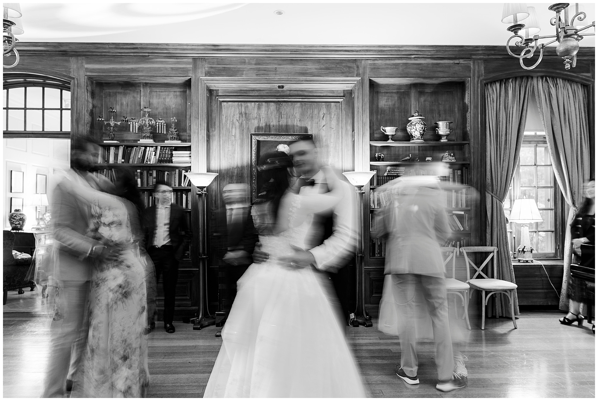 A black-and-white photo shows couples dancing in a wood-paneled room. The image is slightly blurred, capturing the motion of the dancers, while others stand or sit in the background near bookshelves and lamps.