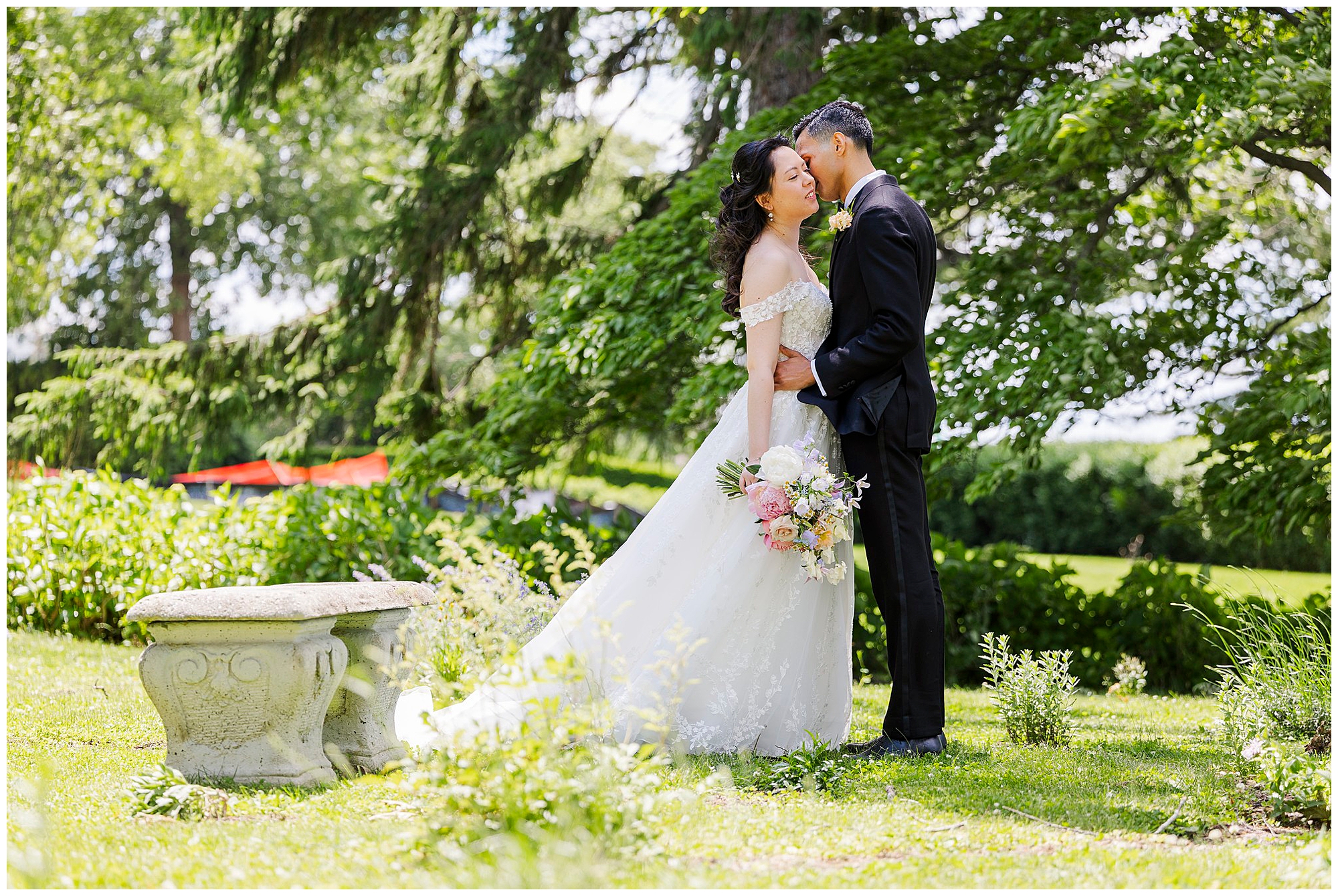 A bride and groom share a joyful moment outdoors, standing close together by a stone bench in a lush garden. The bride holds a bouquet of pink and white flowers, and both are smiling beneath the shade of green trees.