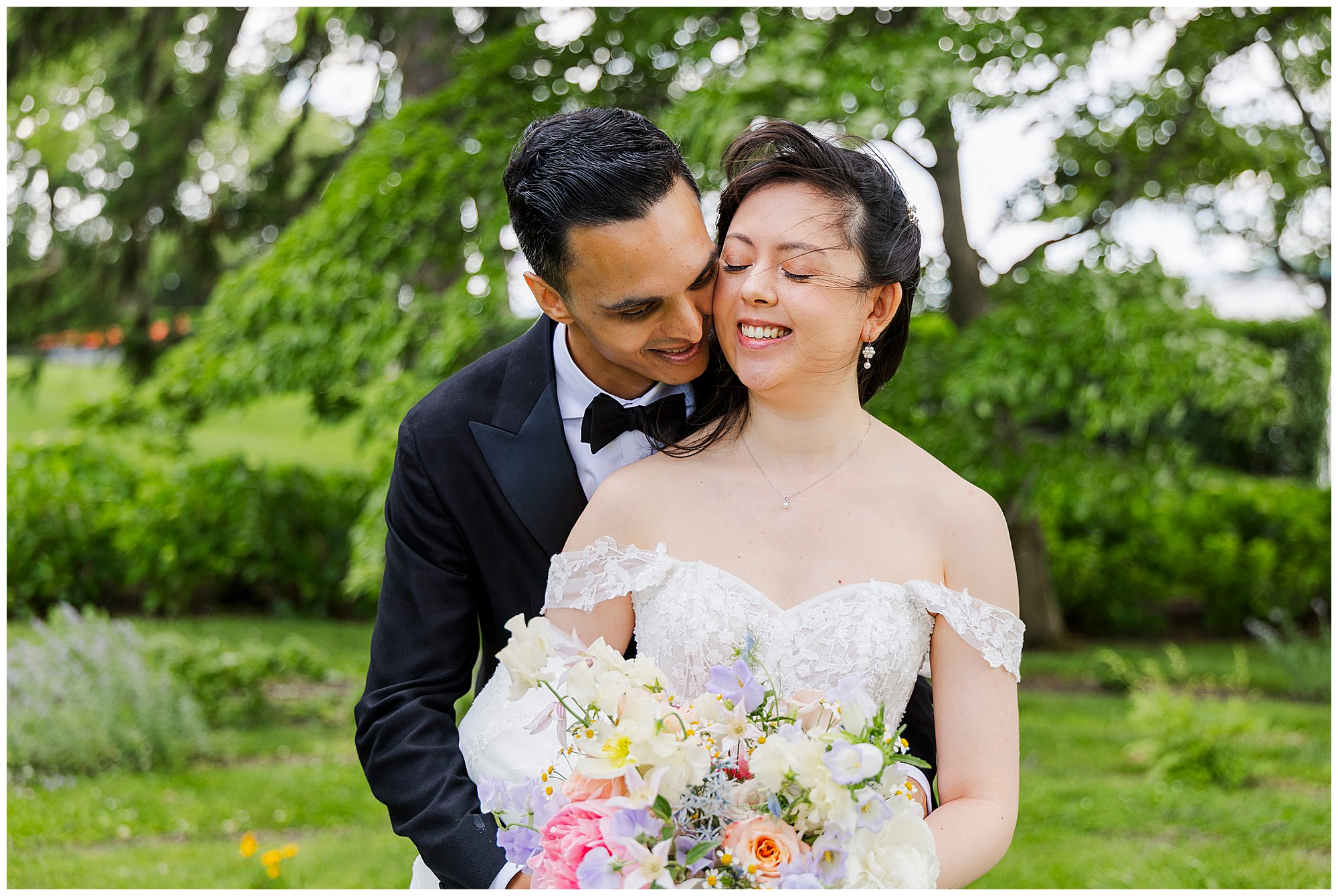 A groom in a black tuxedo lovingly hugs a smiling bride in an off-shoulder white dress holding a colorful bouquet, with lush green trees and grass in the background.