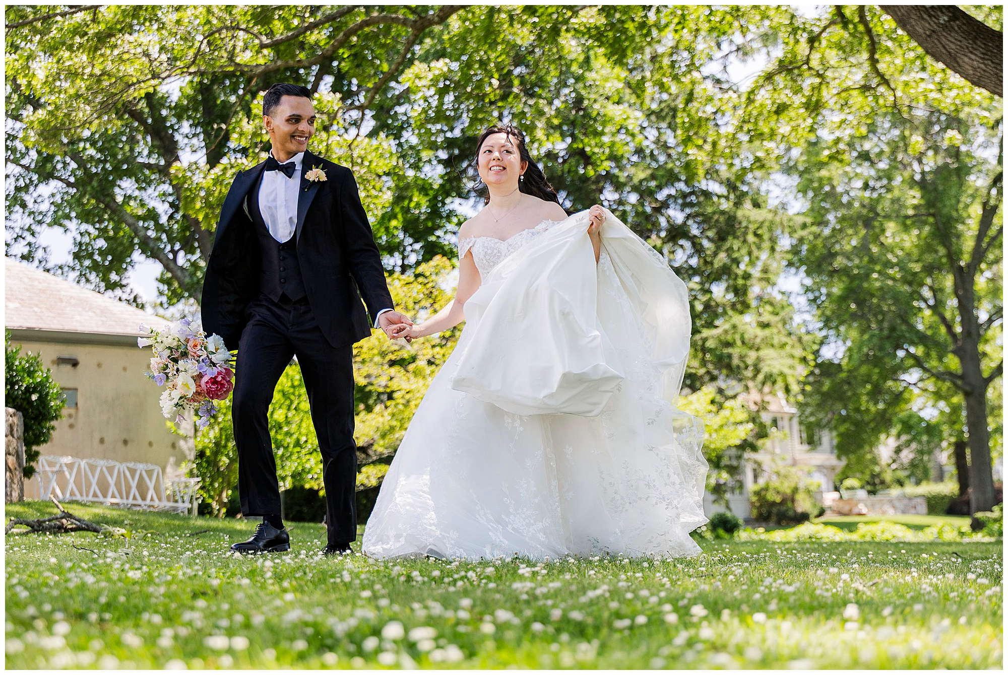 A joyful bride and groom walk hand in hand on a sunny, grassy lawn surrounded by trees. The bride holds her dress and a bouquet, both are smiling and dressed in formal wedding attire.