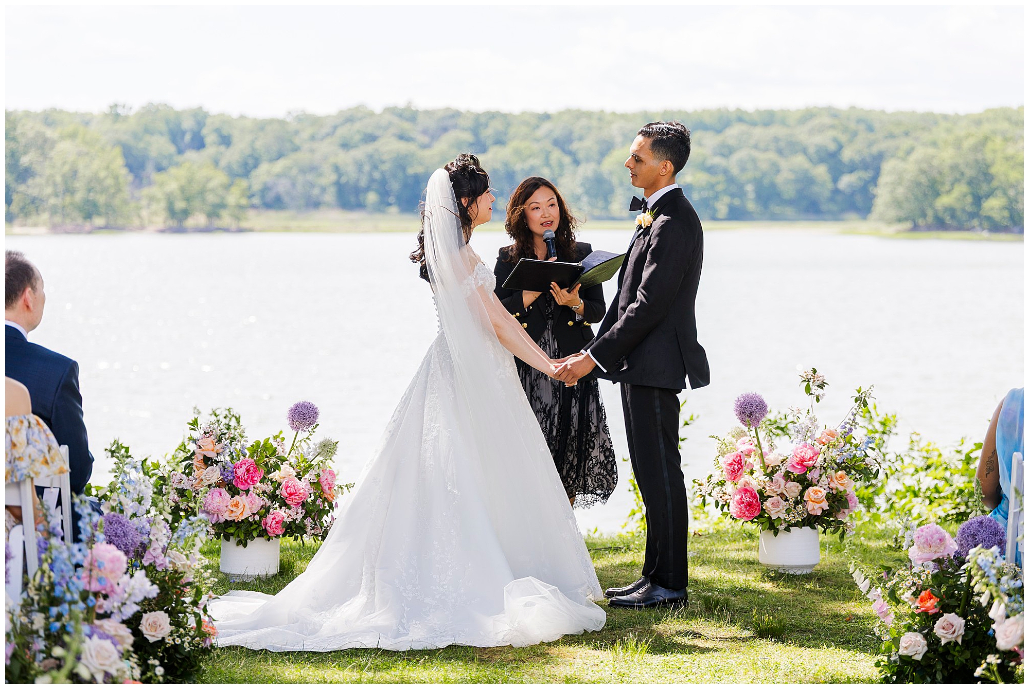 A bride and groom hold hands during their outdoor wedding ceremony by a lake, surrounded by colorful flower arrangements, with an officiant standing between them. Trees and water are visible in the background.
