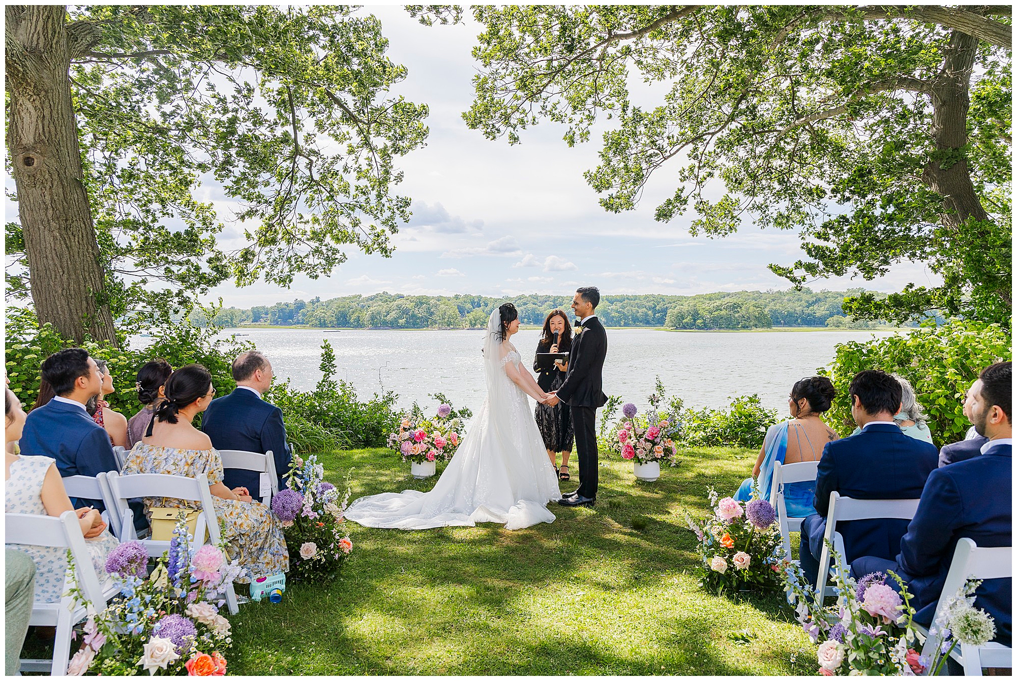 A bride and groom stand facing each other during an outdoor wedding ceremony by a lake, surrounded by guests seated on either side and colorful floral arrangements under large leafy trees.
