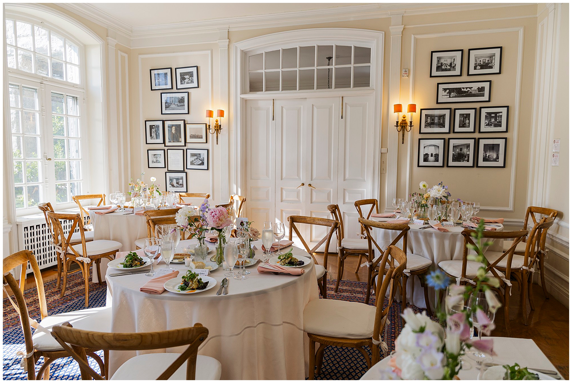 Elegant dining room set for an event with round tables covered in white cloths, wooden chairs, floral centerpieces, neatly arranged plates of salad, and framed black-and-white photos on cream-colored walls.