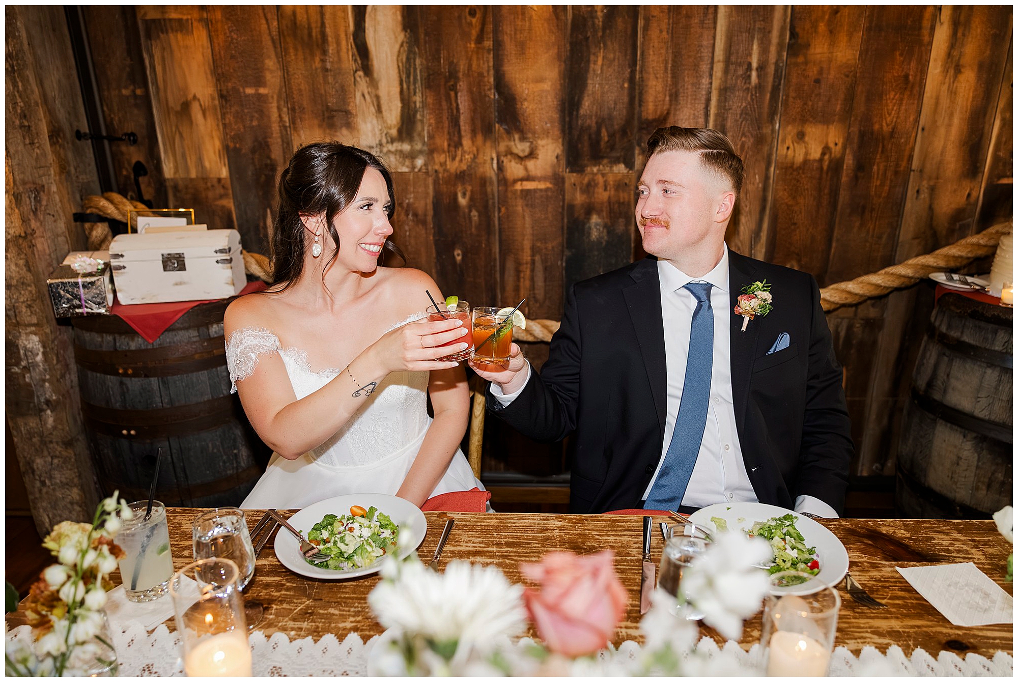 A bride and groom sit at a rustic wooden table, smiling and raising cocktails in a toast. With salads before them and barrels behind, this preston barn wedding exudes a cozy, celebratory atmosphere.