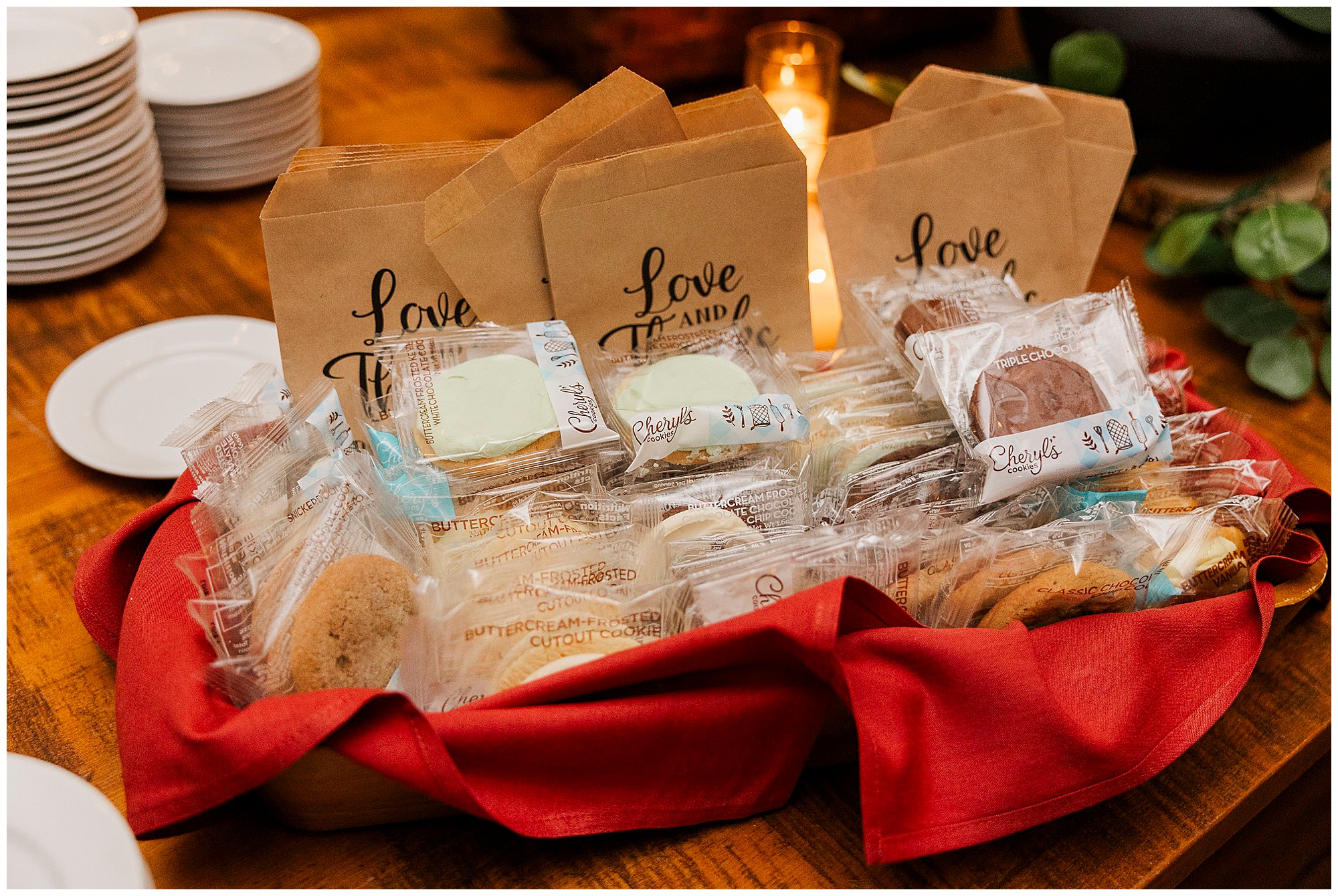 A basket lined with a red cloth holds assorted individually wrapped cookies in front of brown paper bags labeled “Love is Sweet” at a Preston Barn wedding, set on a wooden table with plates and a lit candle in the background.