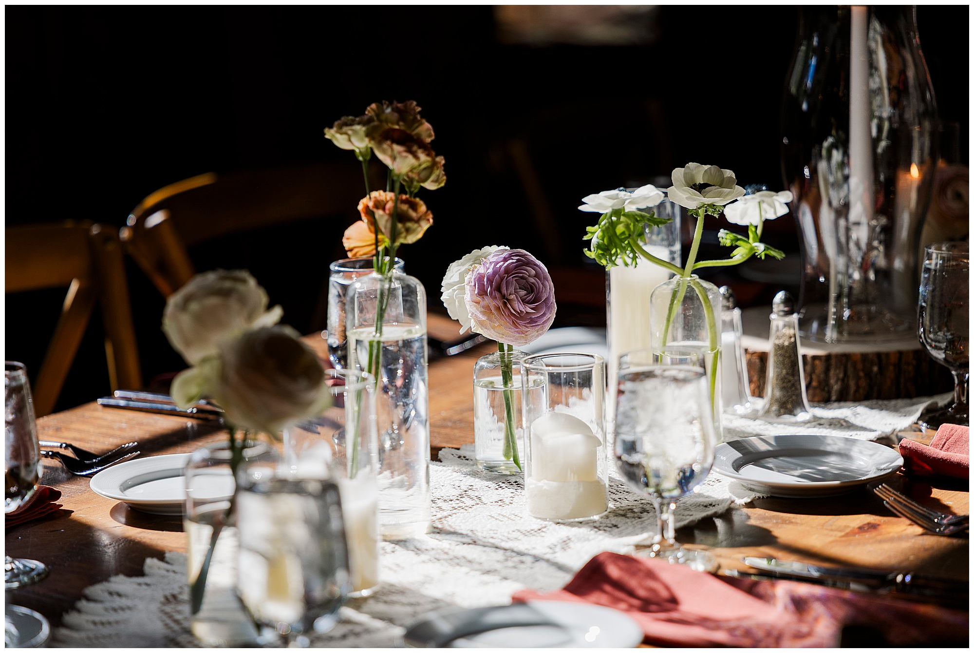 A wooden table set for a meal with glass vases of assorted flowers, glassware, plates, and napkins. Sunlight highlights the centerpiece, creating a warm and inviting atmosphere perfect for a Preston Barn wedding.