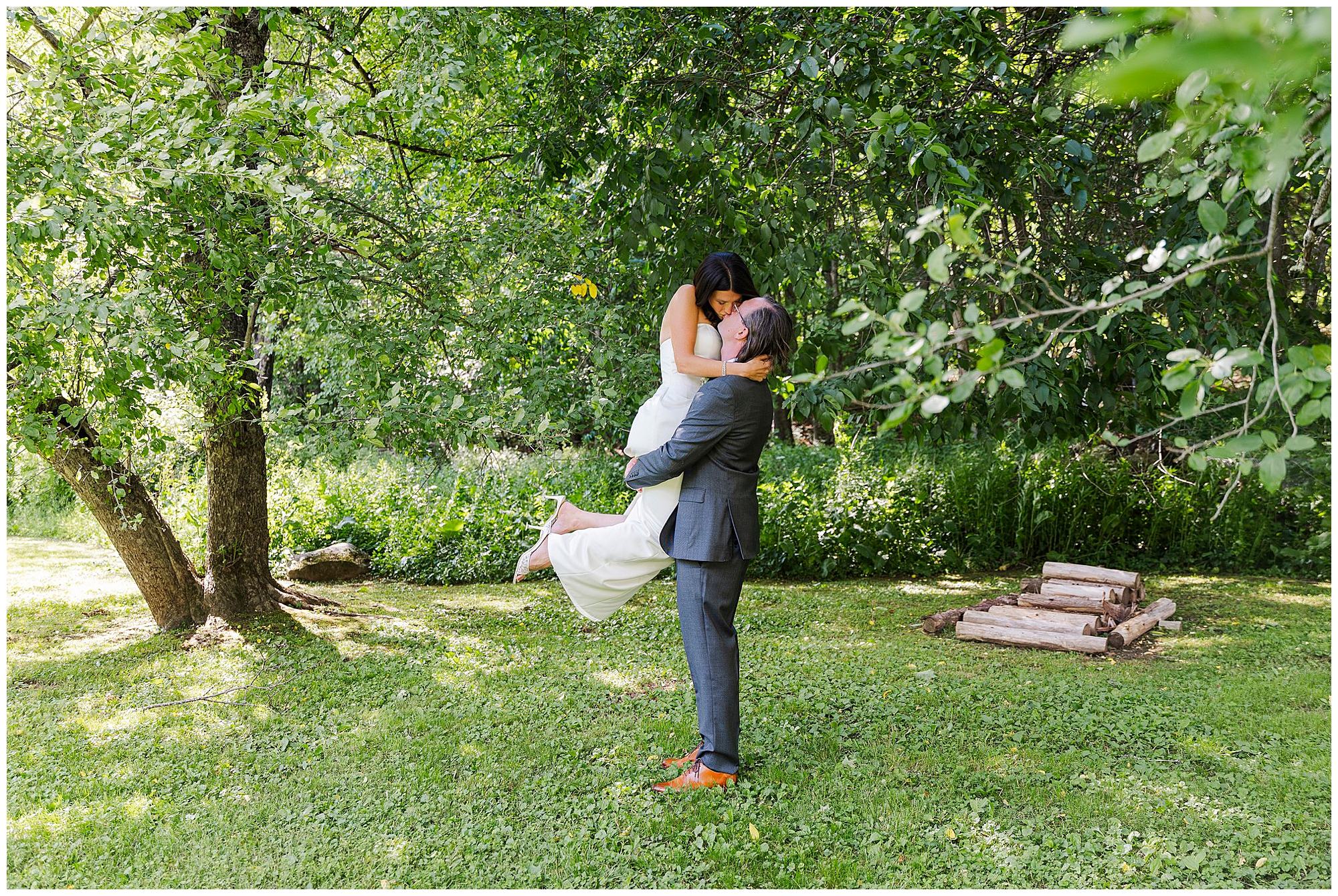 A man in a suit lifts a woman in a white dress, both smiling, under green trees at an intimate outdoor airbnb micro wedding. Logs are stacked on the grass nearby.