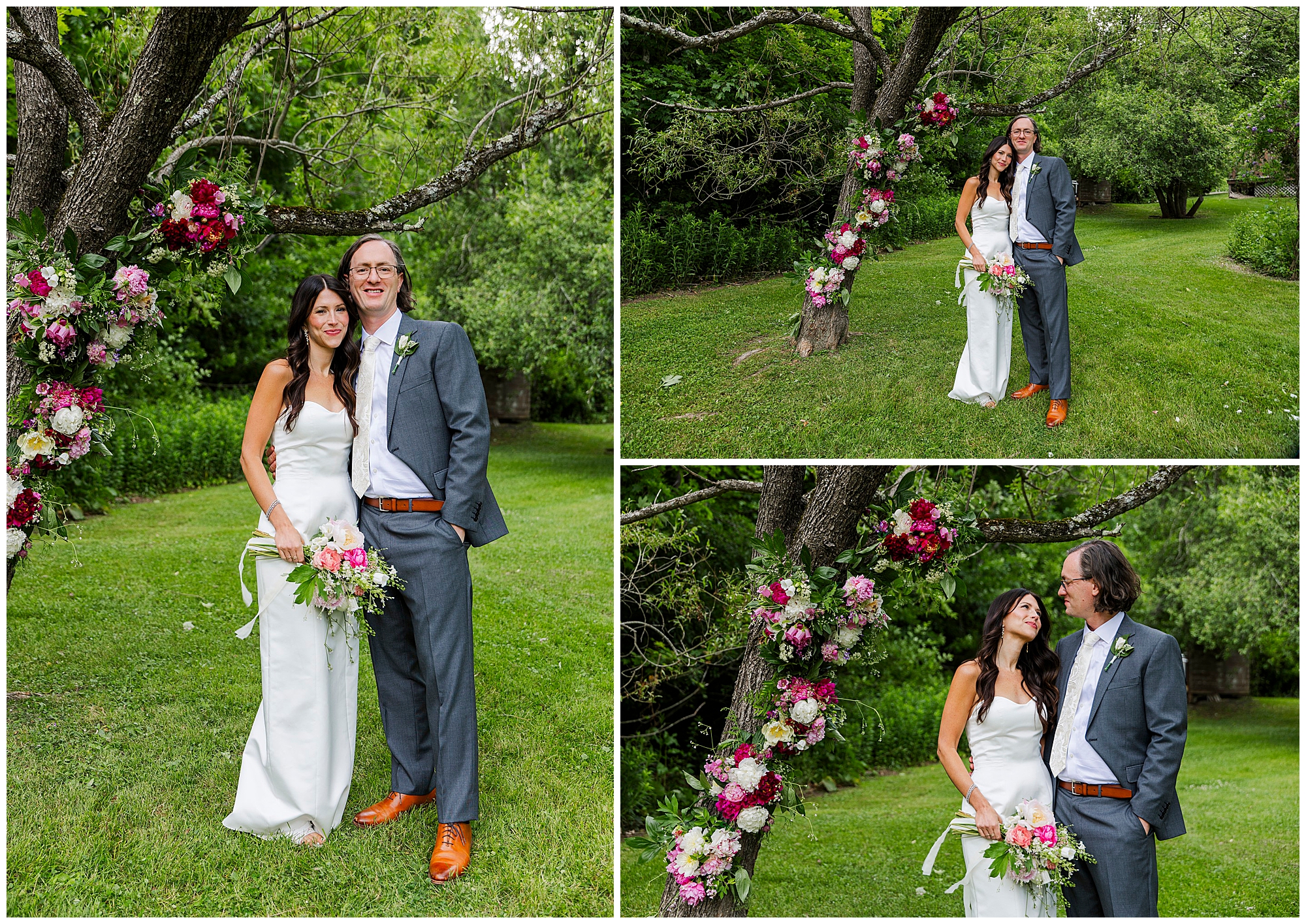 A bride in a white dress and groom in a gray suit stand under a tree decorated with pink and white flowers, smiling and posing together outdoors on the lush green lawn of their intimate Airbnb micro wedding.
