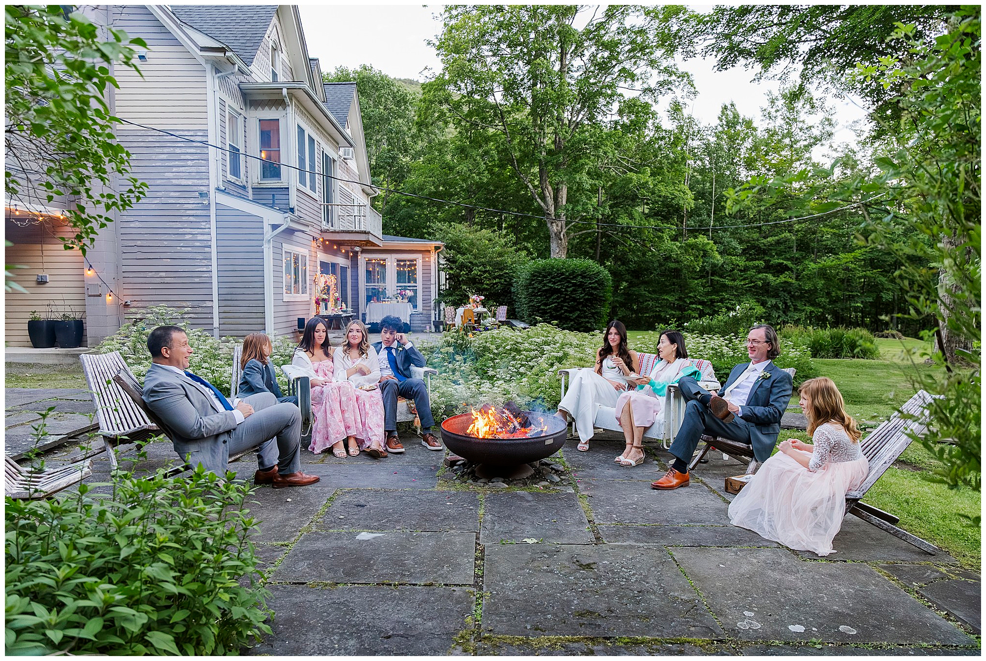 Nine people in formal attire sit in a circle around a fire pit on a stone patio outside a large house, surrounded by greenery—an intimate Airbnb micro wedding on a warm summer evening.
