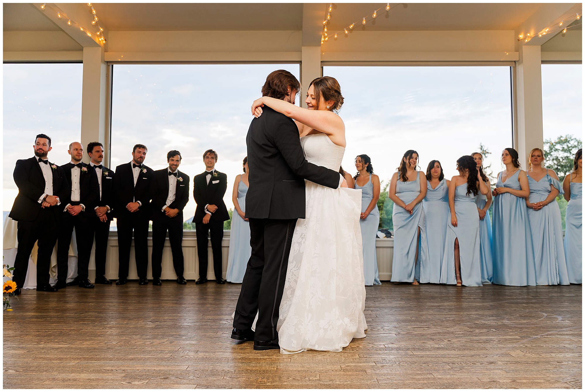 A bride and groom share their first dance in a bright room at a summer garrison wedding, surrounded by bridesmaids in light blue dresses and groomsmen in black tuxedos, all watching them and smiling.