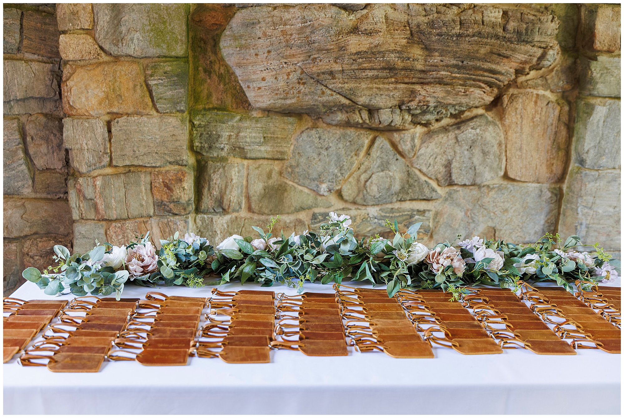 A table with rows of brown leather-bound items, possibly place cards or small notebooks, is set against a stone wall and decorated with a lush green and blush floral arrangement for a stunning wedding at Arrow Park.
