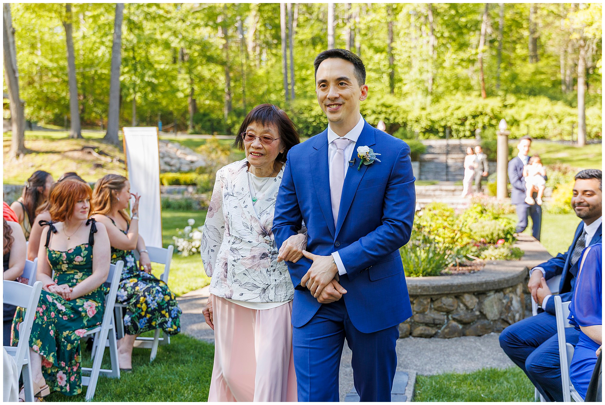 A smiling man in a blue suit walks arm-in-arm with an older woman in a light floral outfit down an outdoor aisle during a wedding at Arrow Park, surrounded by seated guests in a lush garden setting.