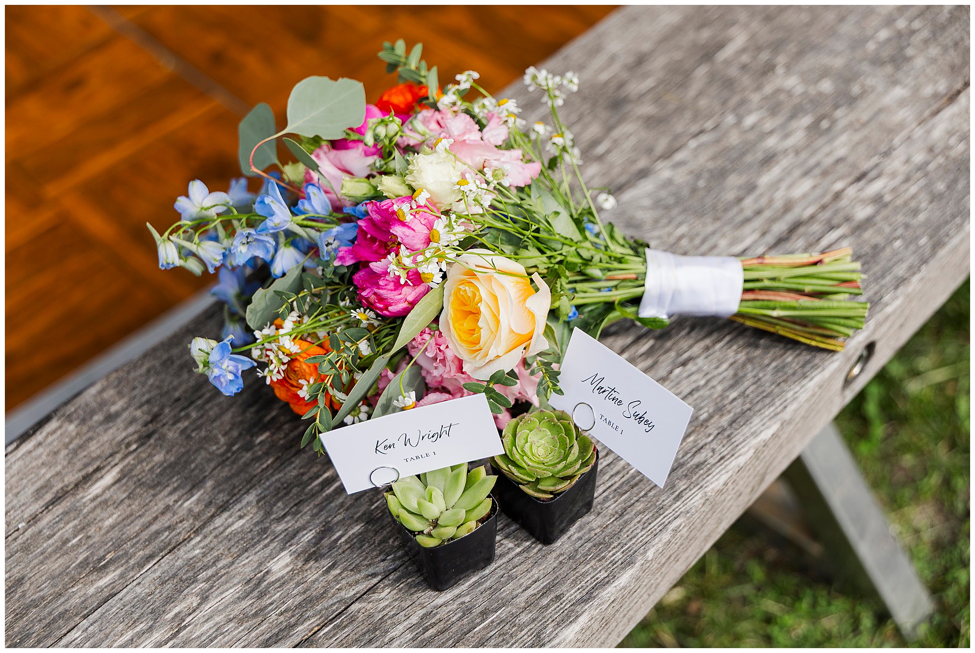 A colorful bouquet of fresh flowers lies on a wooden bench next to two small potted succulents with name cards reading Ken Wright and Marcia Casey, perfect details for a charming wedding at the yard.