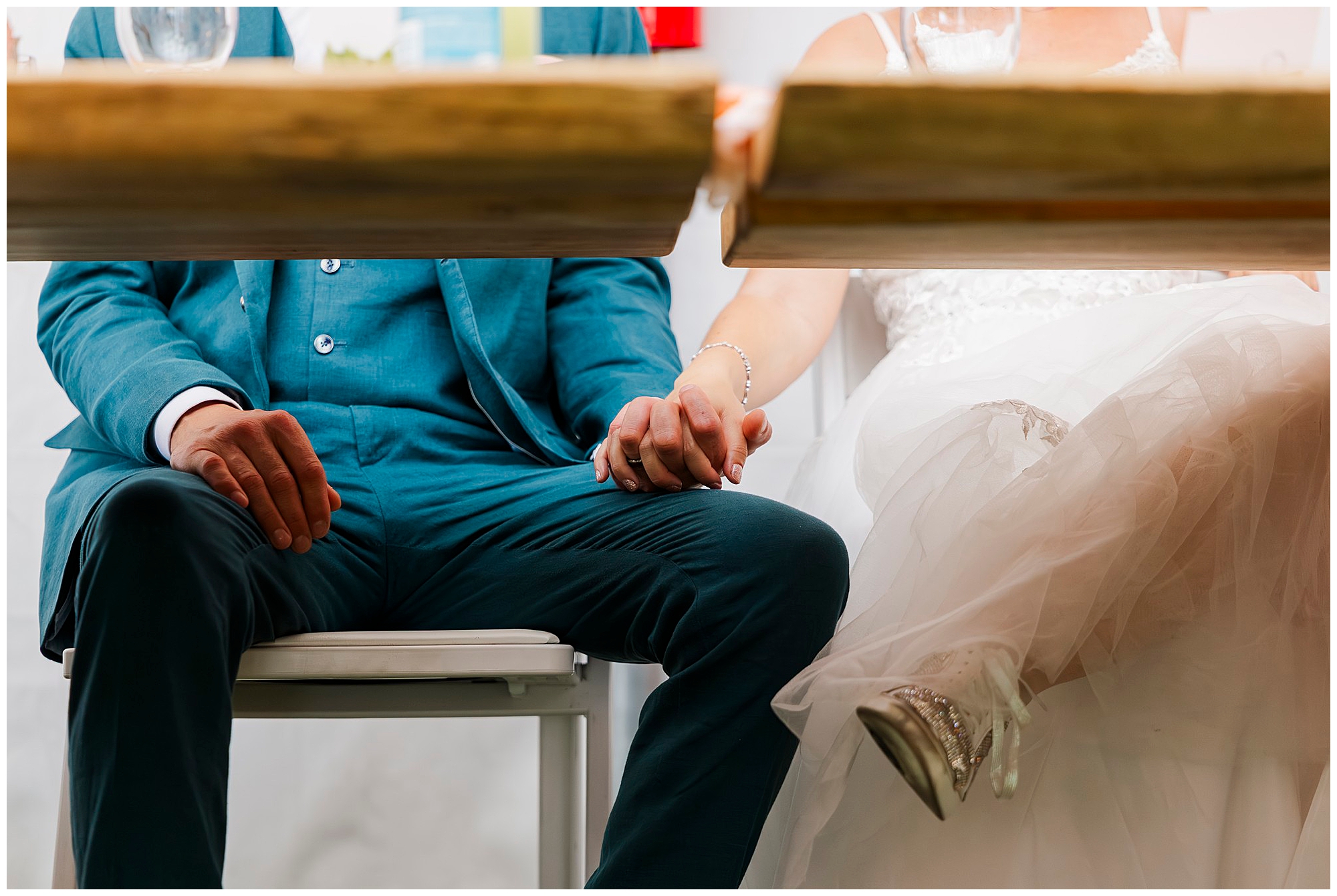 A bride in a white dress and a groom in a teal suit are sitting side by side at their wedding at the yard, holding hands under the table, with only their lower bodies and clasped hands visible.