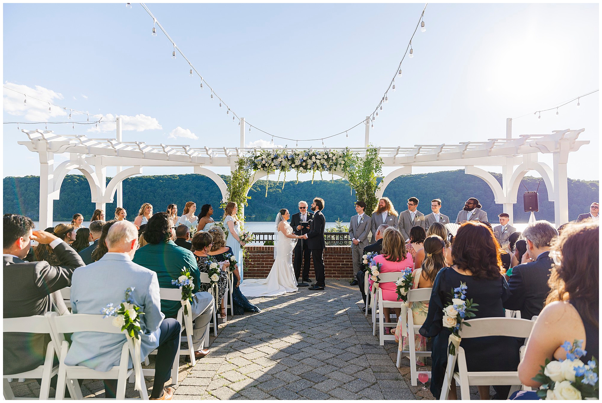 A wedding at The Grandview unfolds outdoors on a sunny day, with a couple standing under a decorated white pergola, surrounded by their wedding party and guests seated in white chairs adorned with flowers.