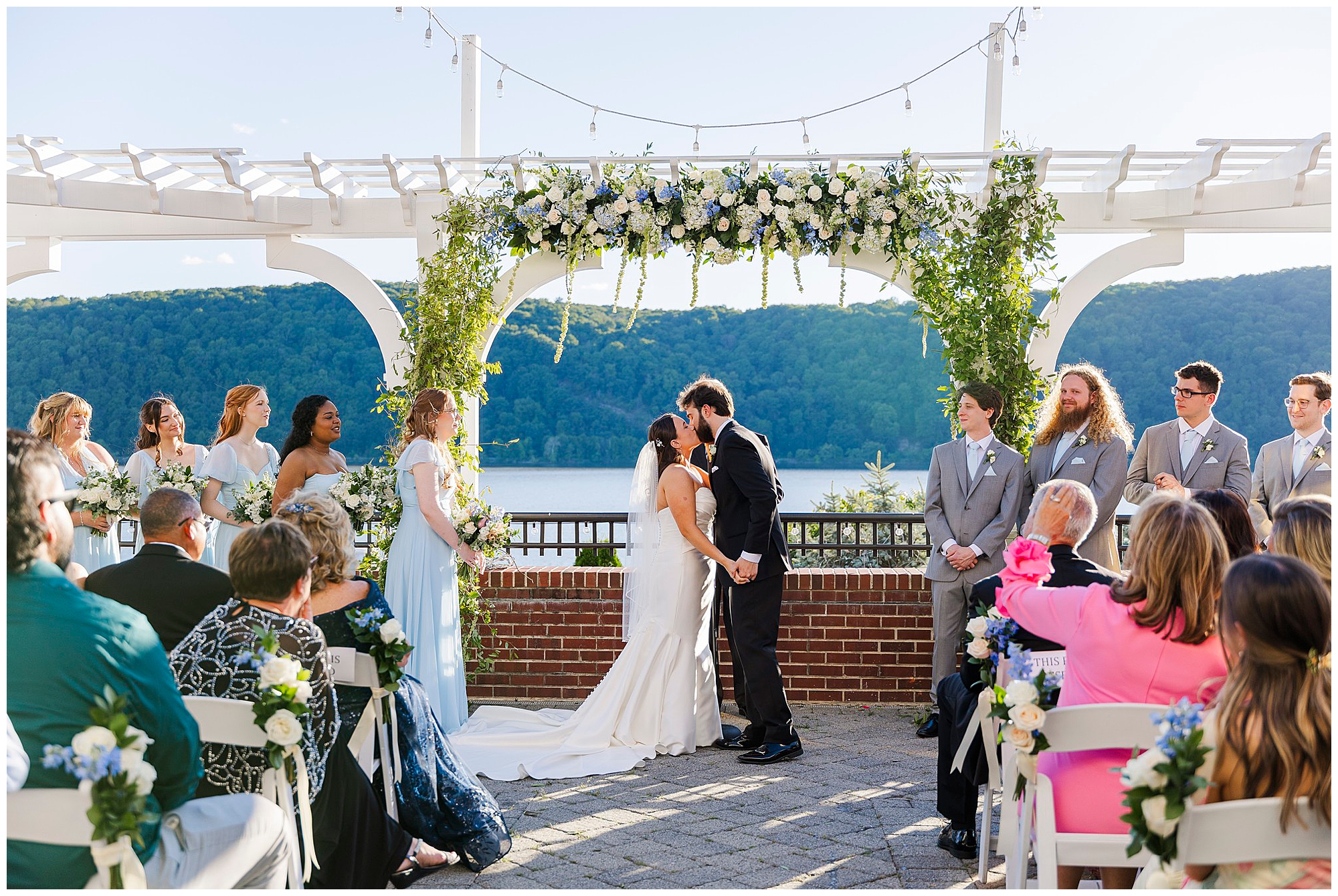 A bride and groom kiss at their wedding at The Grandview, beneath a white floral arbor, surrounded by friends and loved ones, with a scenic lake and green hills creating a stunning backdrop.