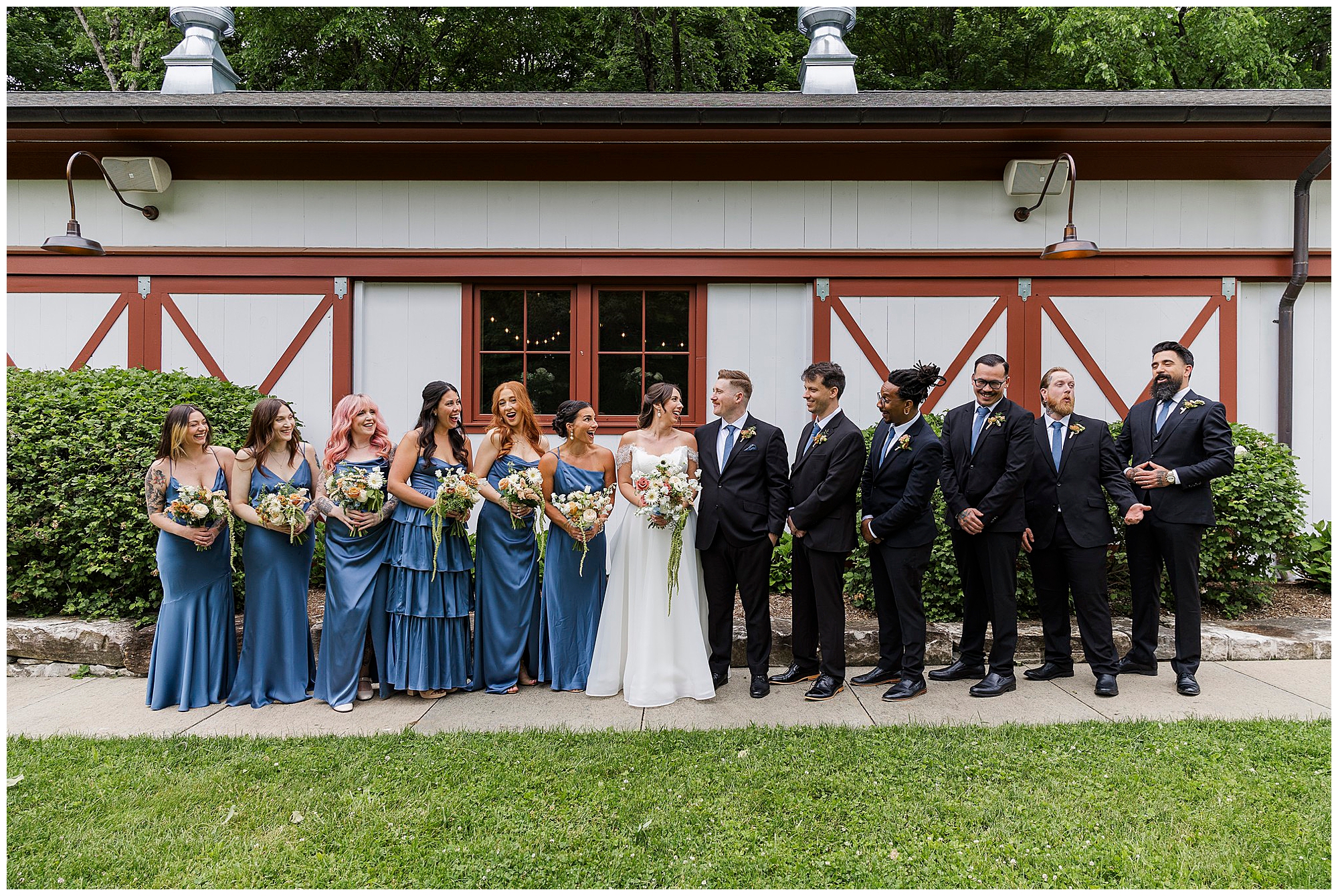 A wedding party stands in a row outside Preston Barn with red trim. Bridesmaids in blue dresses hold bouquets, while groomsmen in black suits stand beside the bride and groom, all smiling and laughing together at this unforgettable Preston Barn wedding.
