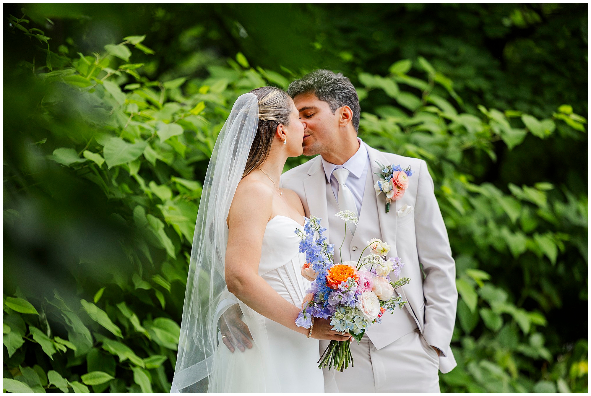 A bride and groom share a kiss outdoors, surrounded by lush greenery during their wedding photos at the Roundhouse. The bride holds a colorful bouquet, her white dress and veil complementing the groom’s light suit and floral boutonniere.