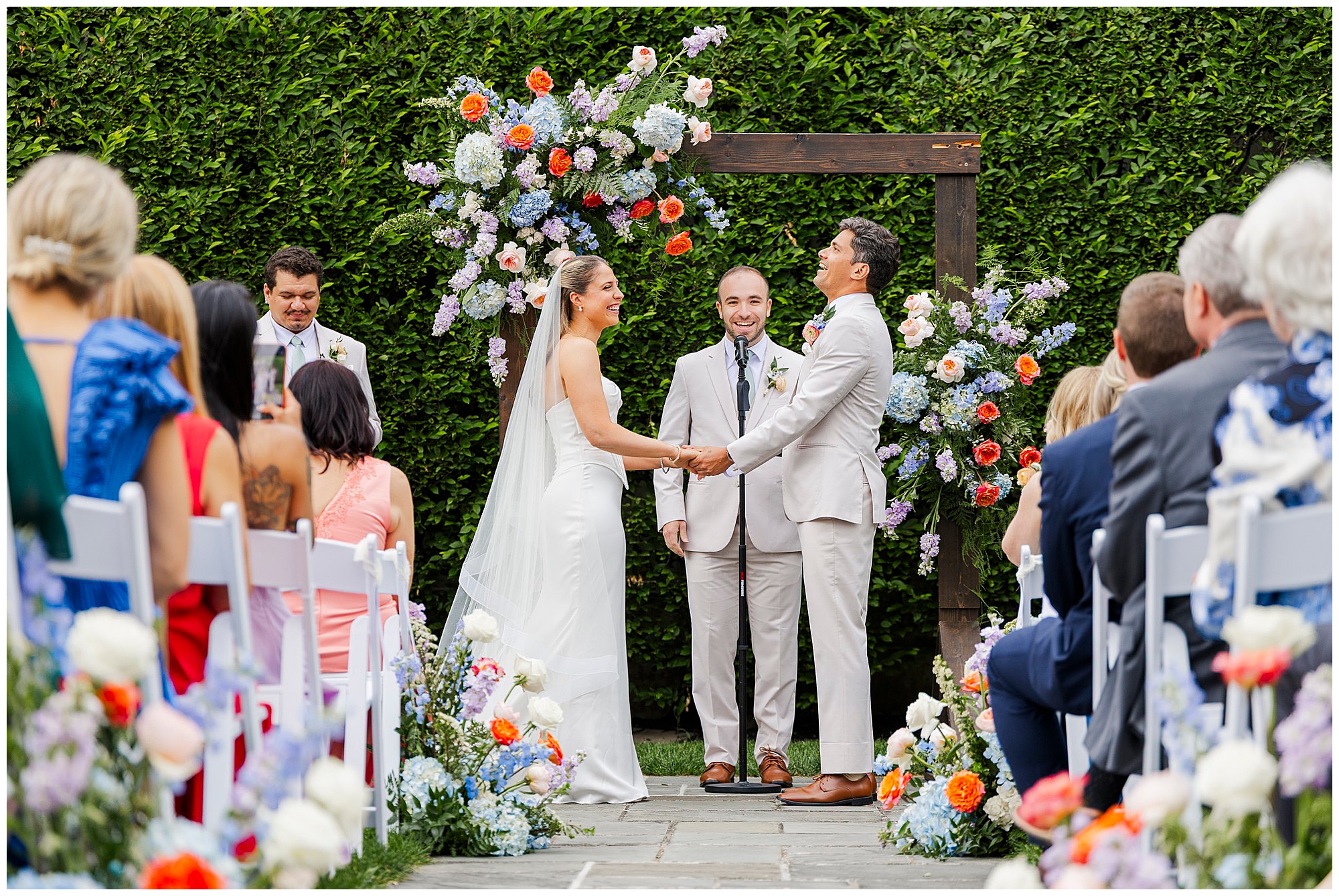 A bride and groom stand under a floral arch, holding hands and smiling during an outdoor wedding ceremony at the Roundhouse, surrounded by seated guests and colorful flower arrangements—perfect for unforgettable wedding photos at the Roundhouse.