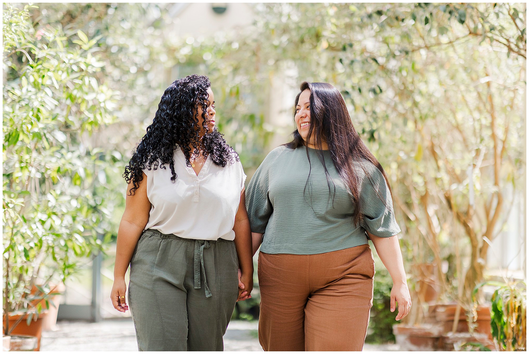 Two women holding hands and smiling at each other while walking outdoors on a sunny day at Stonecrop Gardens, surrounded by green plants and trees—capturing joyful LGBTQ engagement photos.