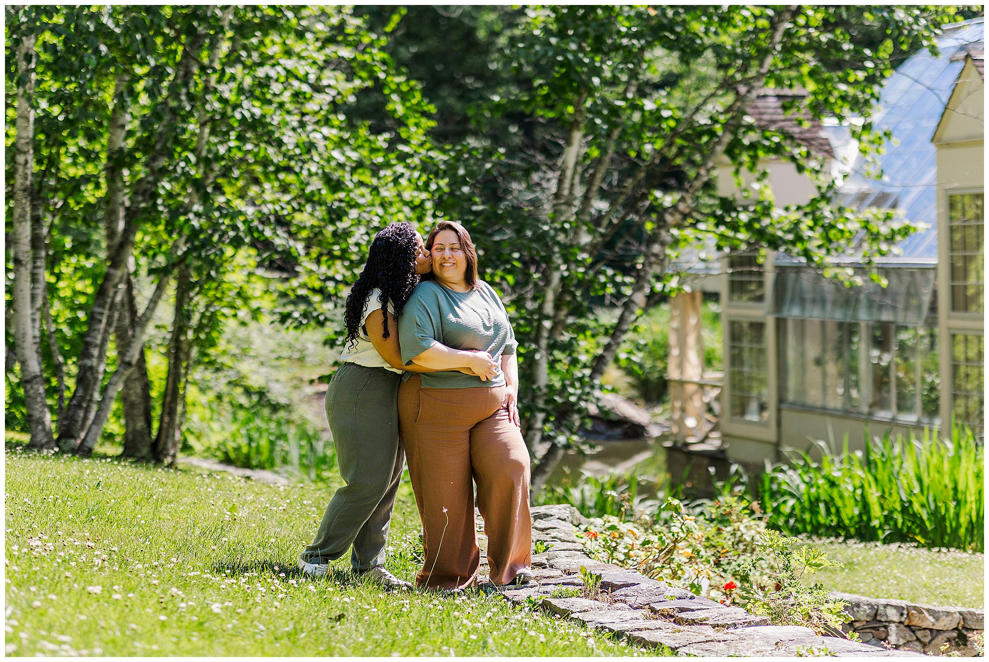 Two people stand on a grassy lawn by a stone border at Stonecrop Gardens. One hugs and kisses the other’s cheek as they both smile, capturing joyful LGBTQ engagement photos with trees and a glass greenhouse in the bright sunlight.