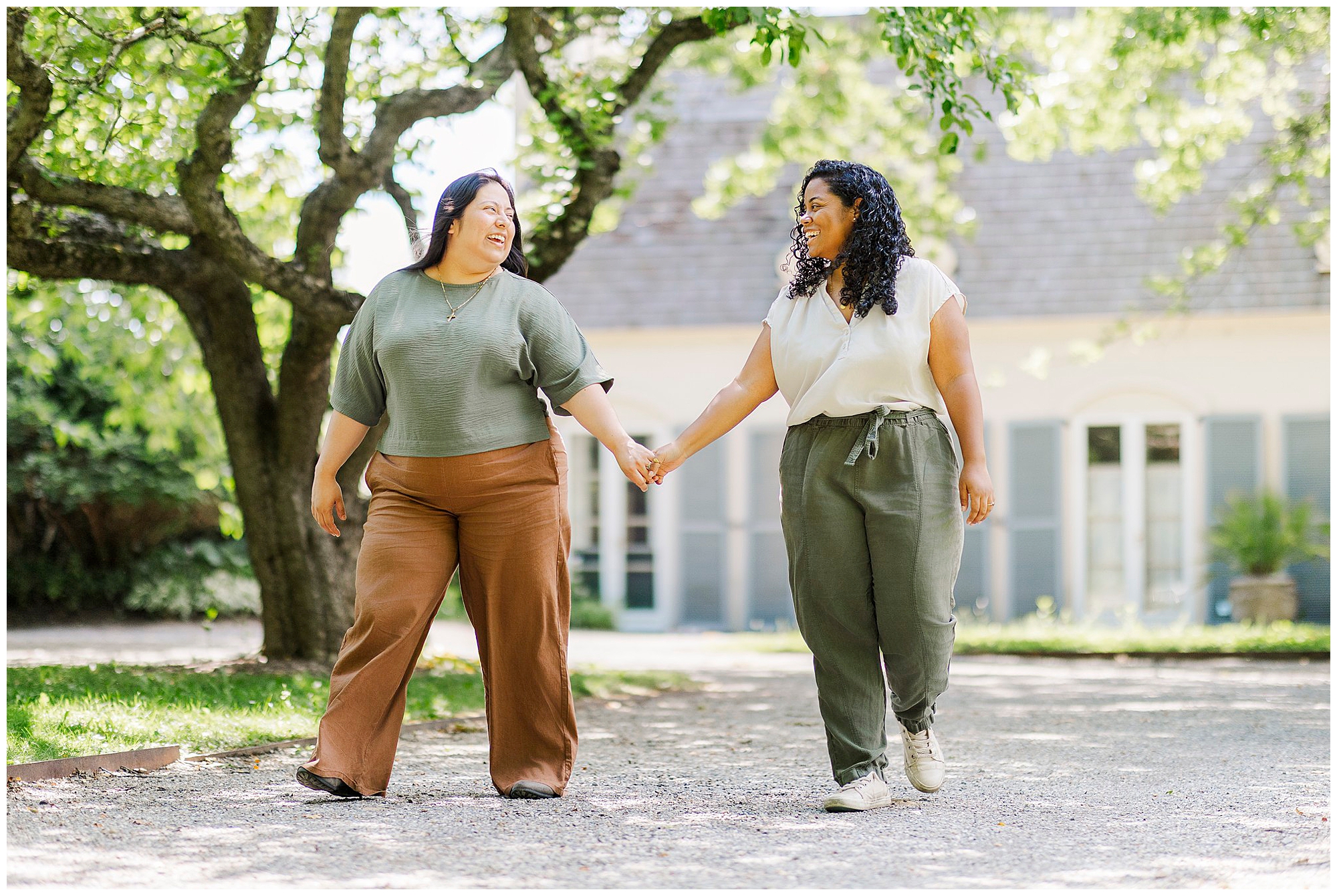 Two women walk hand in hand outside in a sunlit garden, smiling at each other. Captured as LGBTQ engagement photos at Stonecrop Gardens, they are dressed casually and surrounded by trees and greenery, with a building in the background.