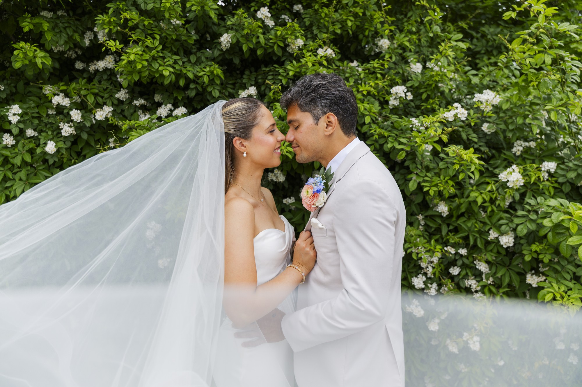 A bride and groom embrace outdoors in front of green bushes with white flowers. The bride’s veil flows to the side, and they gaze at each other, smiling lovingly, both dressed in white formal attire.