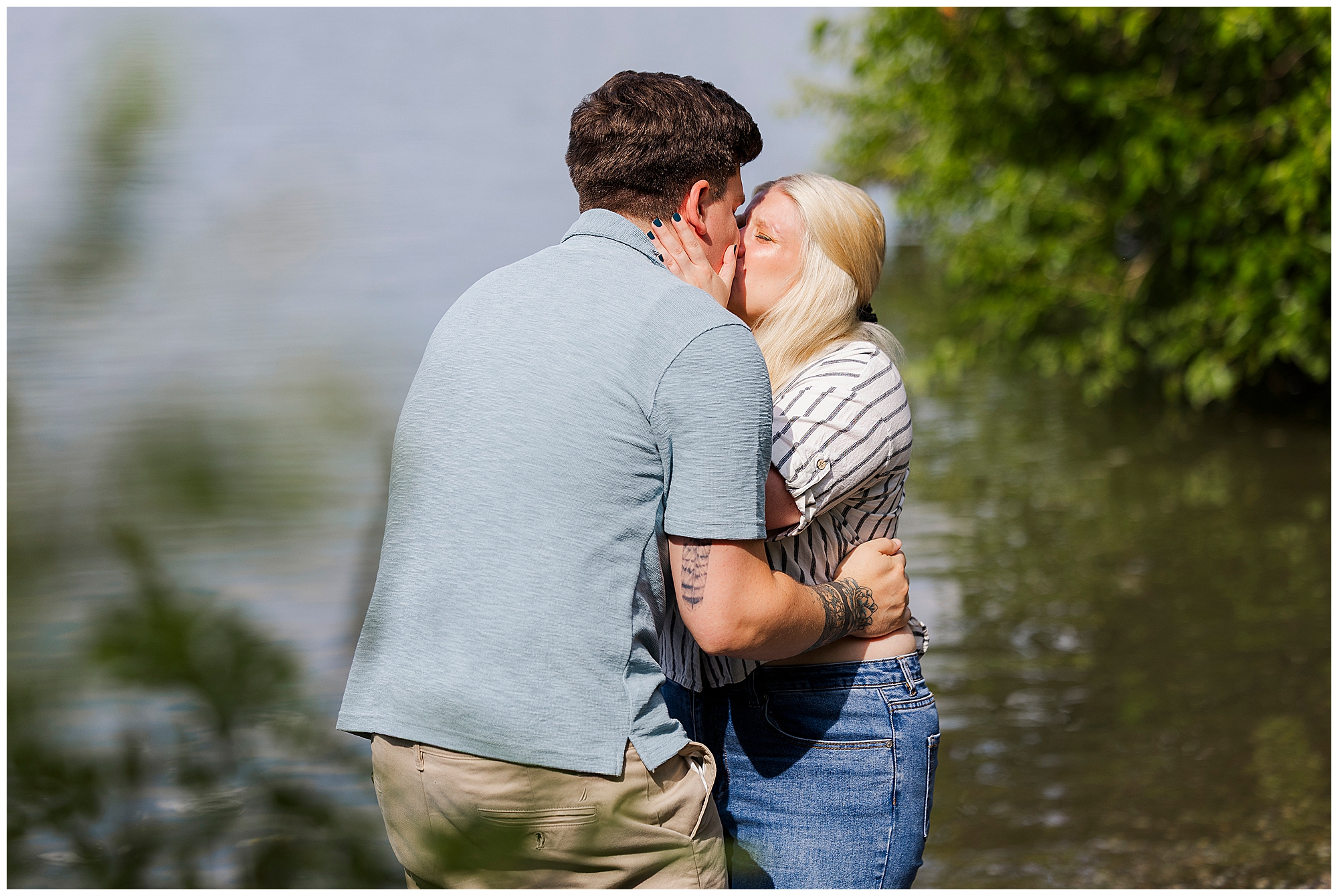 A couple kisses by the water, surrounded by greenery, capturing a romantic moment reminiscent of beach proposal photos. The man wears a light blue shirt and khaki shorts, while the woman wears a striped shirt and jeans.