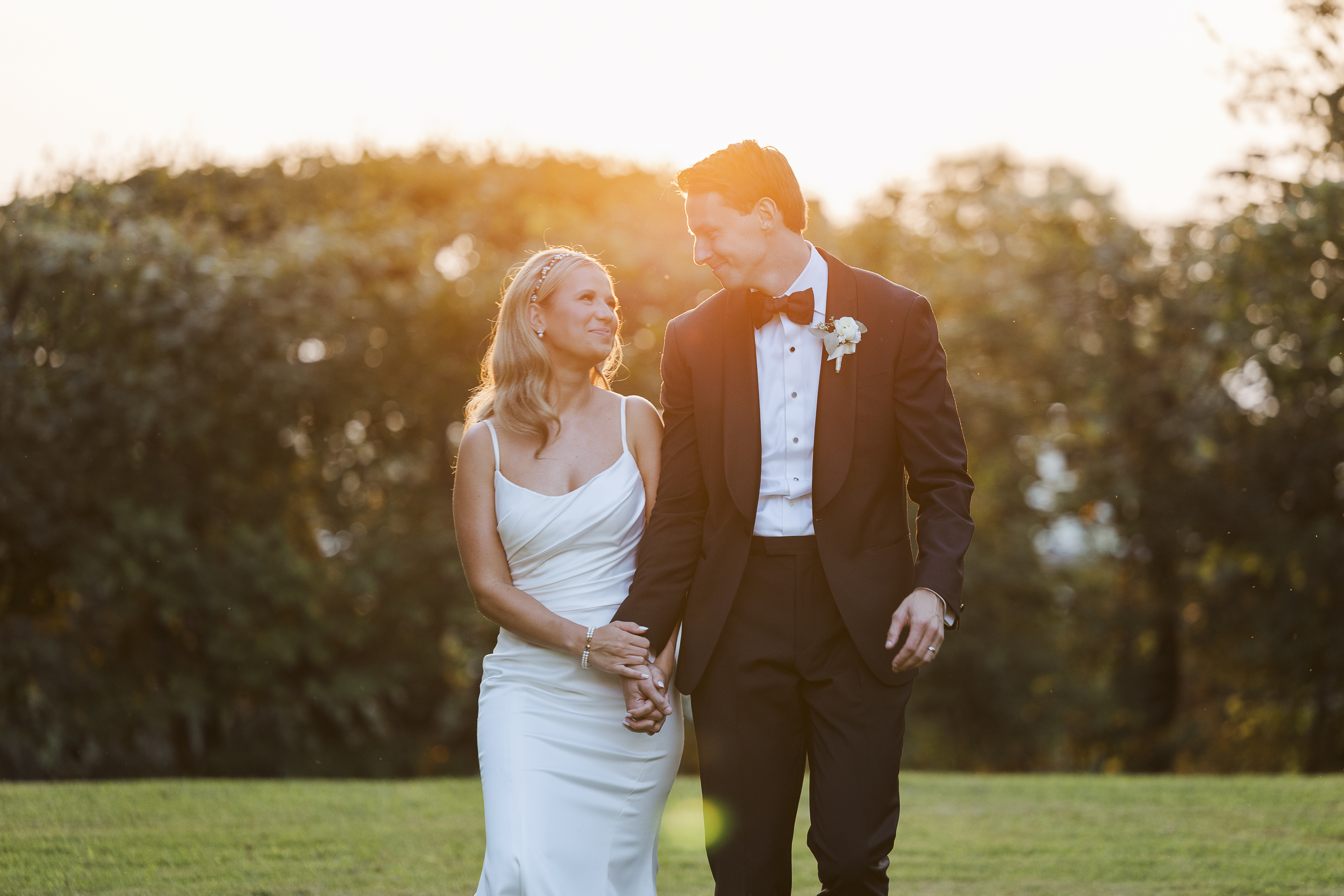 A bride in a white dress and a groom in a black tuxedo walk hand in hand on grass, smiling at each other with sunlight in the background.