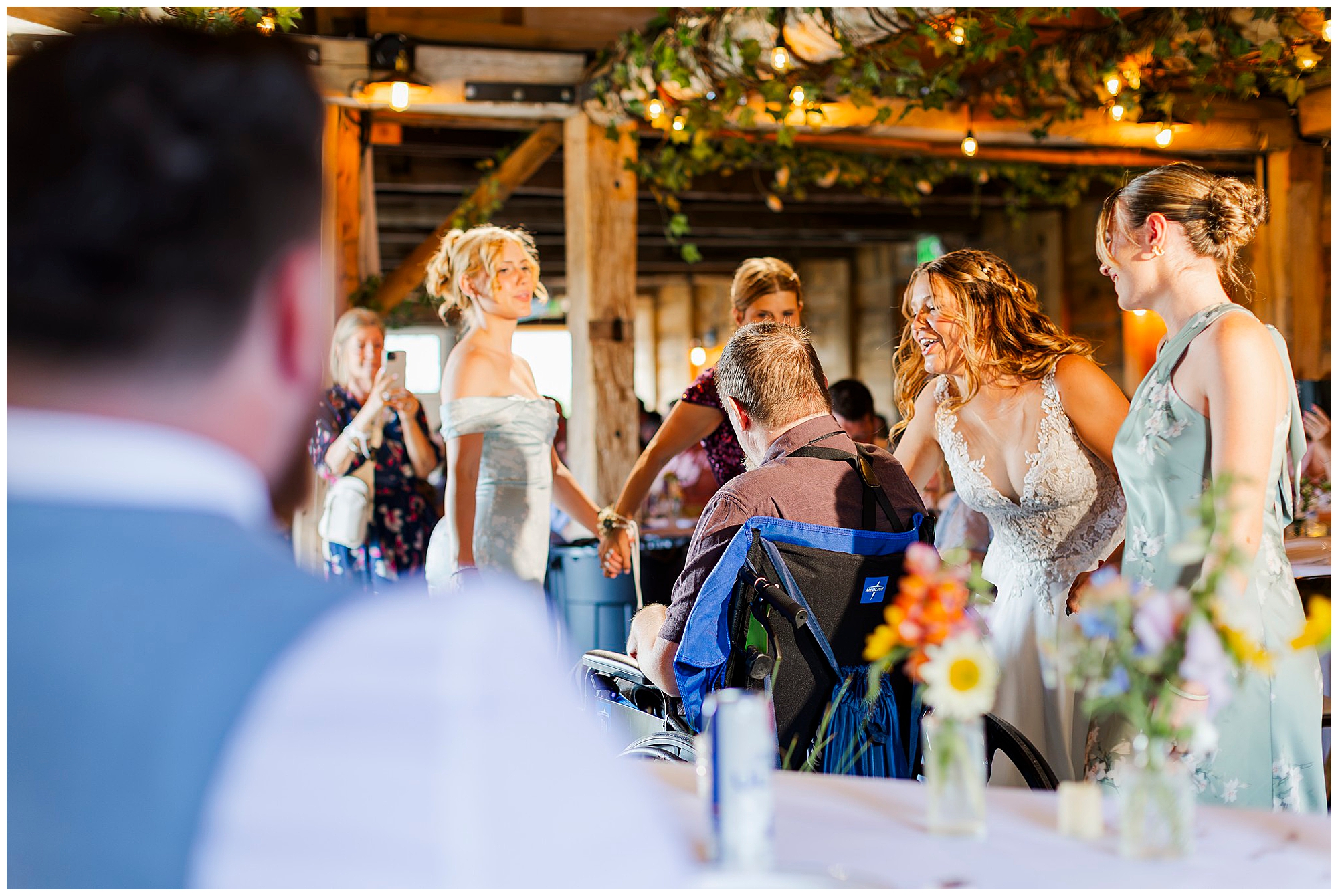 A bride in a white dress smiles and talks with a guest in a wheelchair at a lively indoor knotting hill farm wedding reception, surrounded by bridesmaids in pastel dresses.