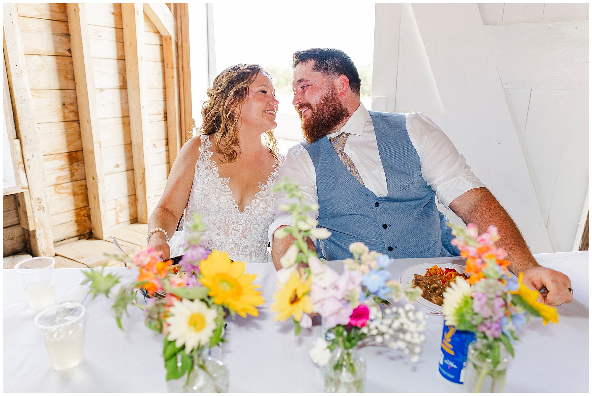 A bride and groom sit at a table decorated with colorful flowers, smiling and looking at each other, in a bright rustic setting during their Knotting Hill Farm wedding reception.