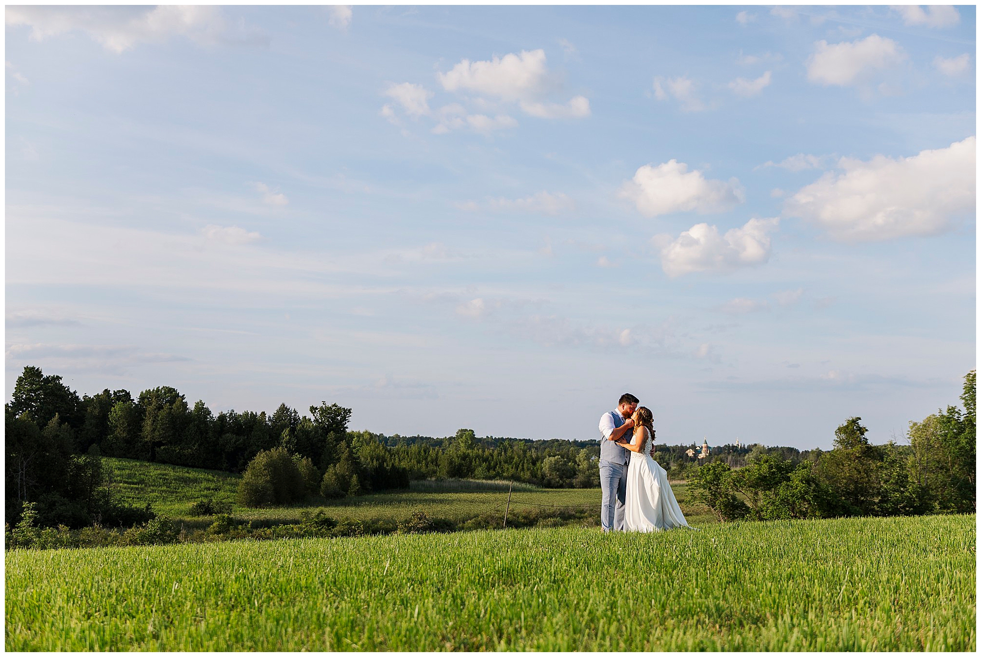 A bride and groom embrace in a green field under a blue sky with scattered clouds at Knotting Hill Farm Wedding, surrounded by trees and distant hills.