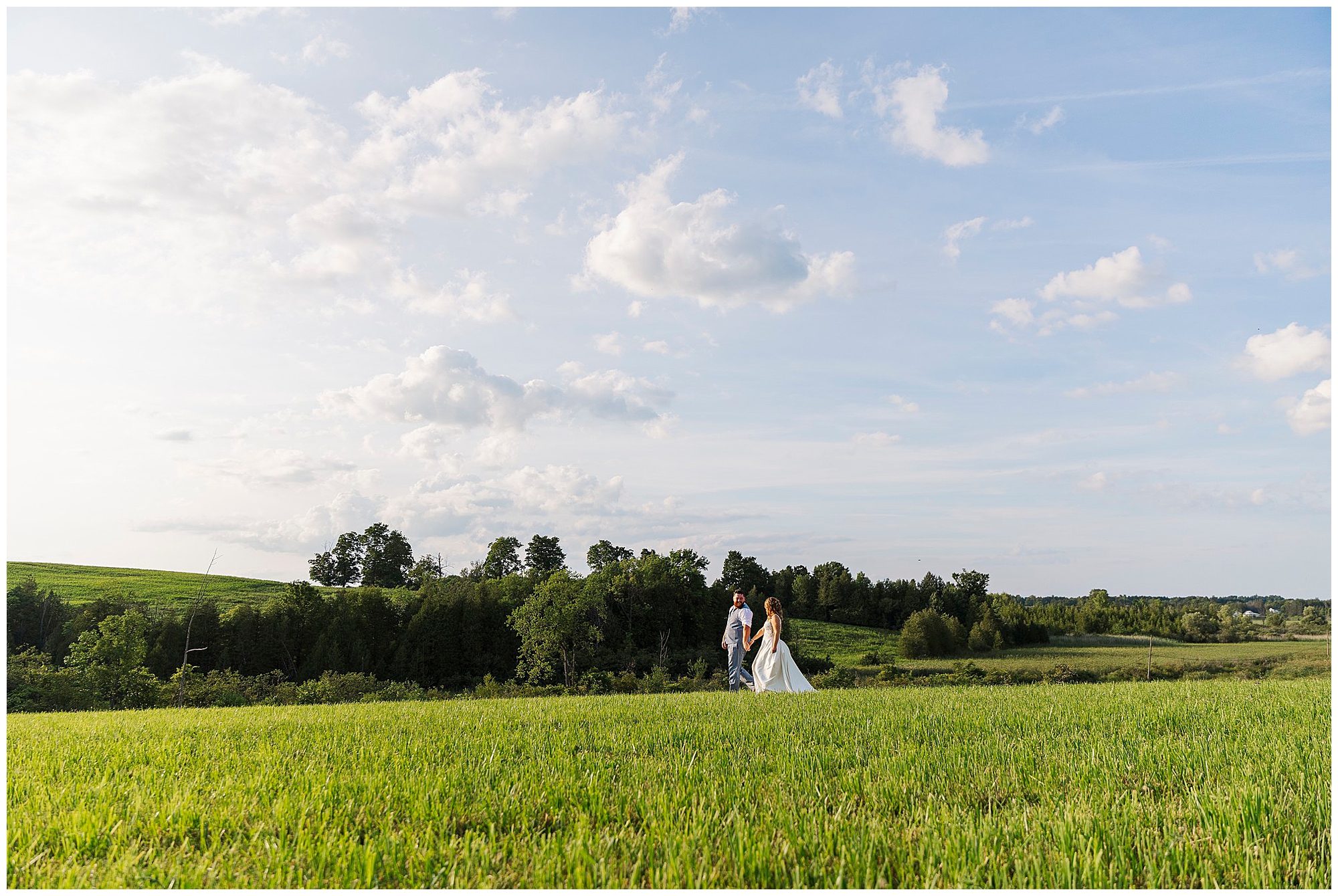 A couple in wedding attire walks hand in hand across a green field under a blue sky with scattered clouds, with trees and rolling hills in the background, capturing the romantic ambiance of a Knotting Hill Farm wedding.