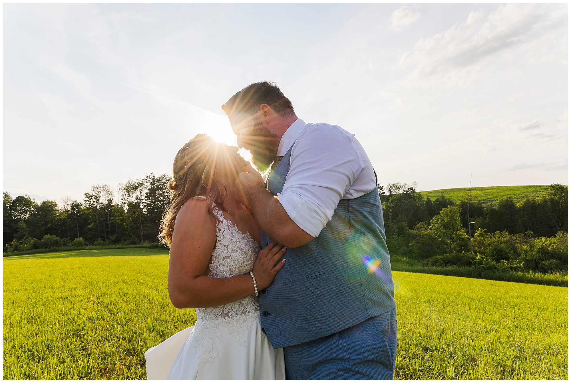 A couple embraces in a sunlit field at sunset during their Knotting Hill Farm wedding, with the sun shining between their faces. The woman wears a white wedding dress, and the man wears a blue vest and tie. Lush greenery and trees fill the background.
