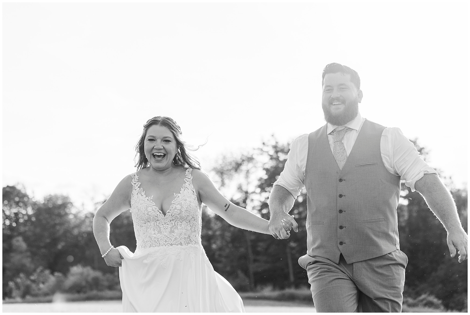 A joyful bride and groom hold hands and run outdoors at Knotting Hill Farm wedding, smiling widely. The bride wears a flowing dress, and the groom is in a vest and tie. Trees and sunlight fill the background.