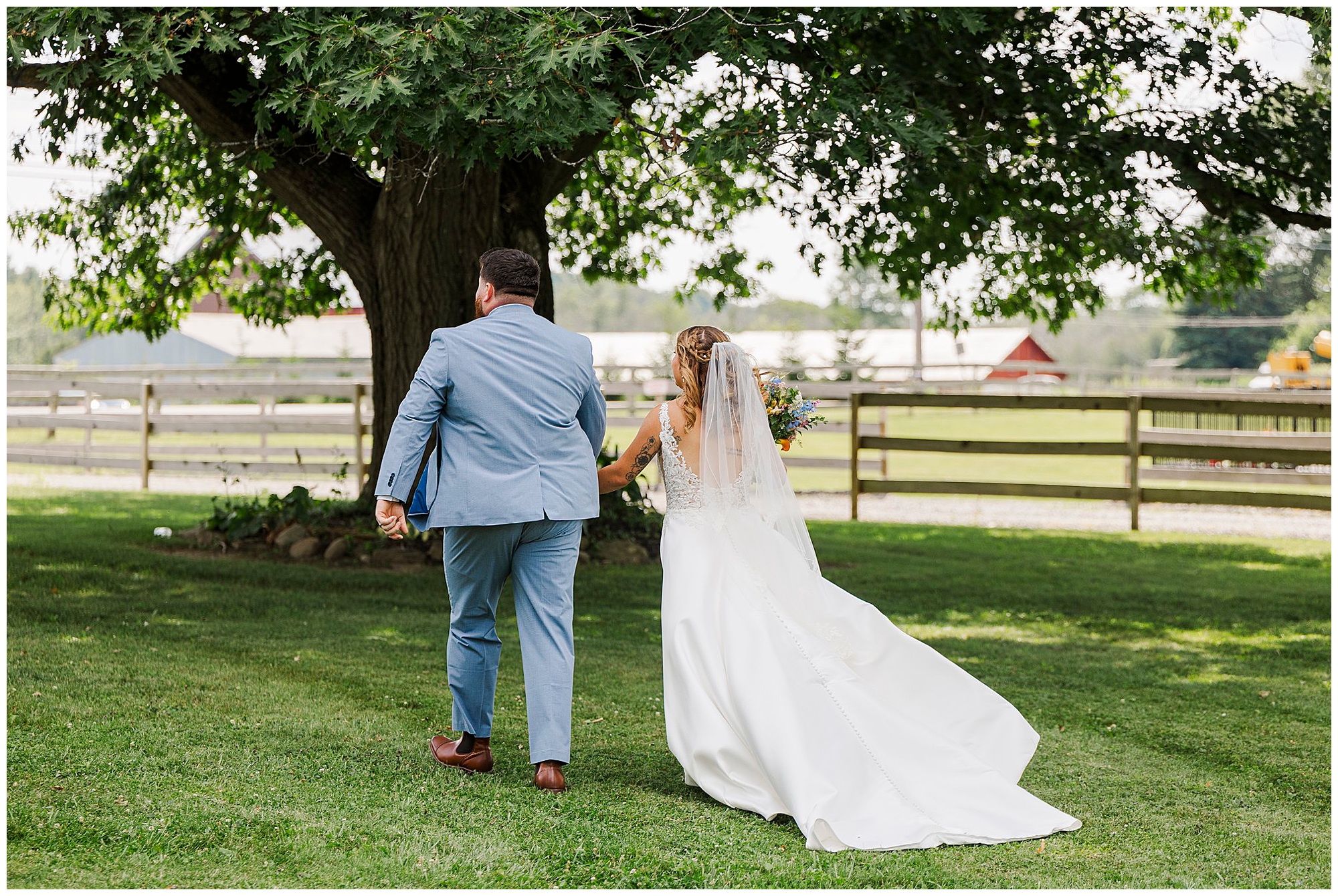 A bride in a white dress and veil walks hand-in-hand with a groom in a light blue suit on green grass under a large tree, with a wooden fence and barns in the background—capturing the rustic charm of a Knotting Hill Farm wedding.