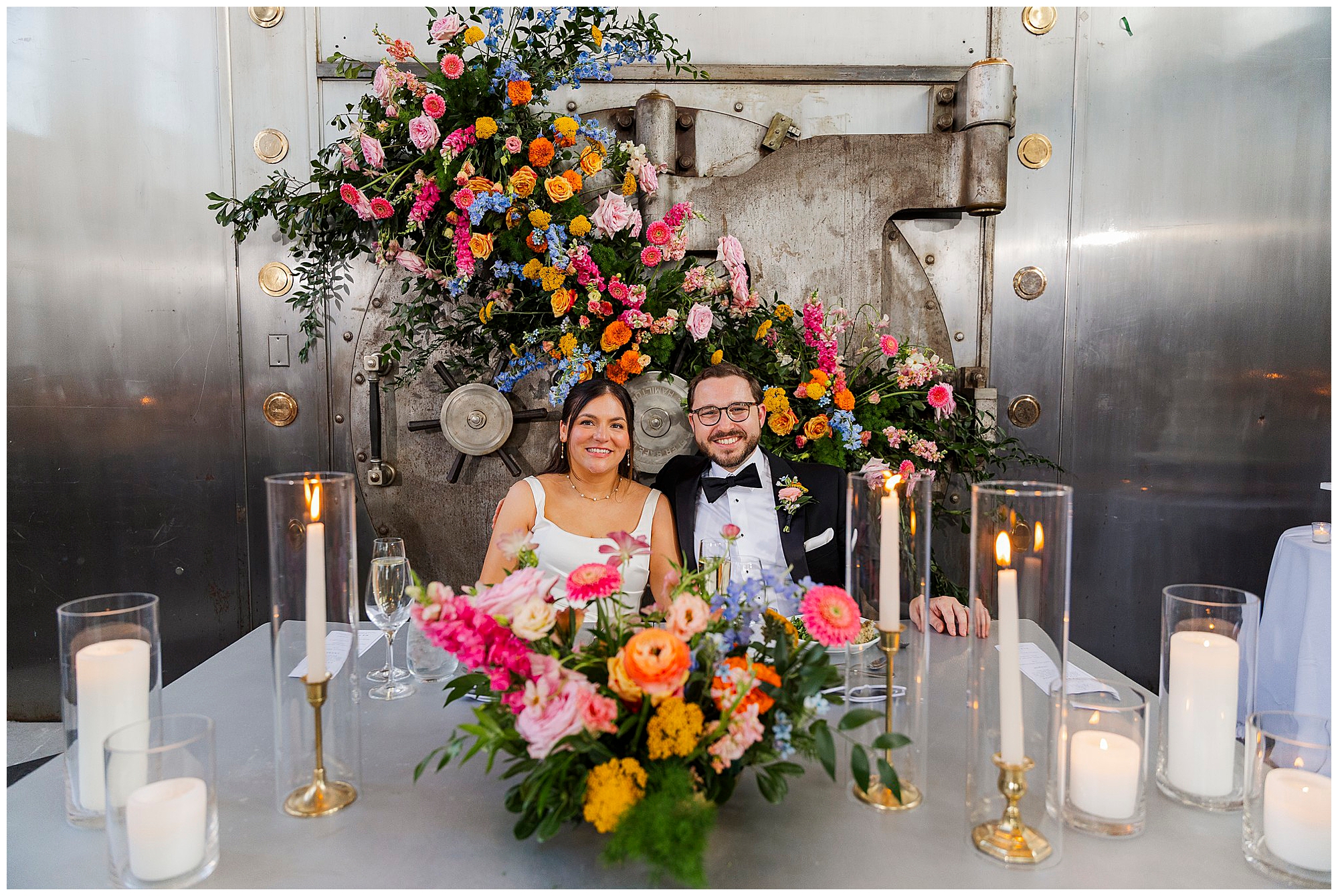 A smiling couple in wedding attire sits at a table with flowers and candles in front of a large metal vault door decorated with colorful floral arrangements, creating a truly unique Felina wedding atmosphere.