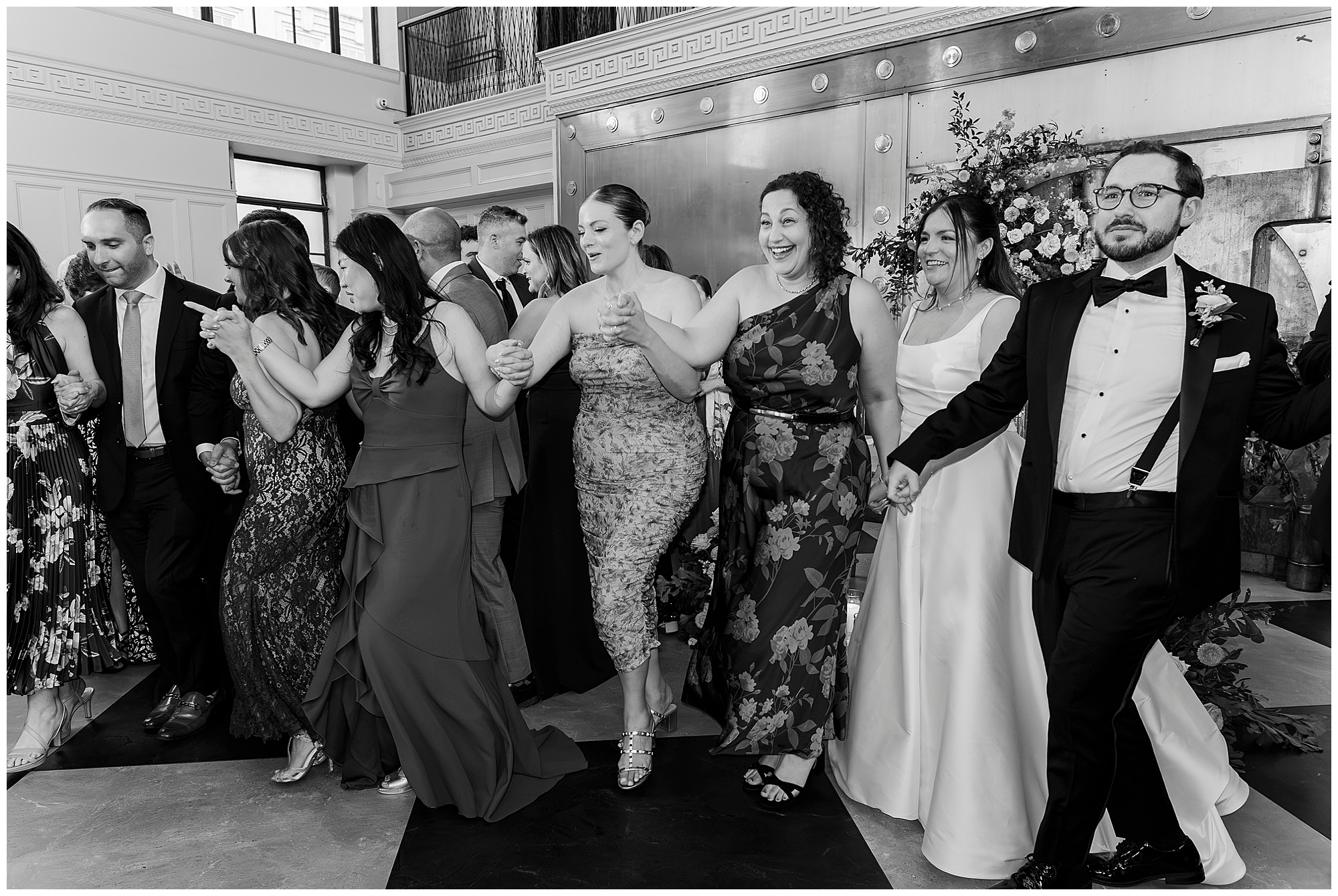 A group of people in formal attire, including a bride in a white dress, hold hands and dance together in a bright, elegant room decorated with flowers at a stunning Felina wedding.