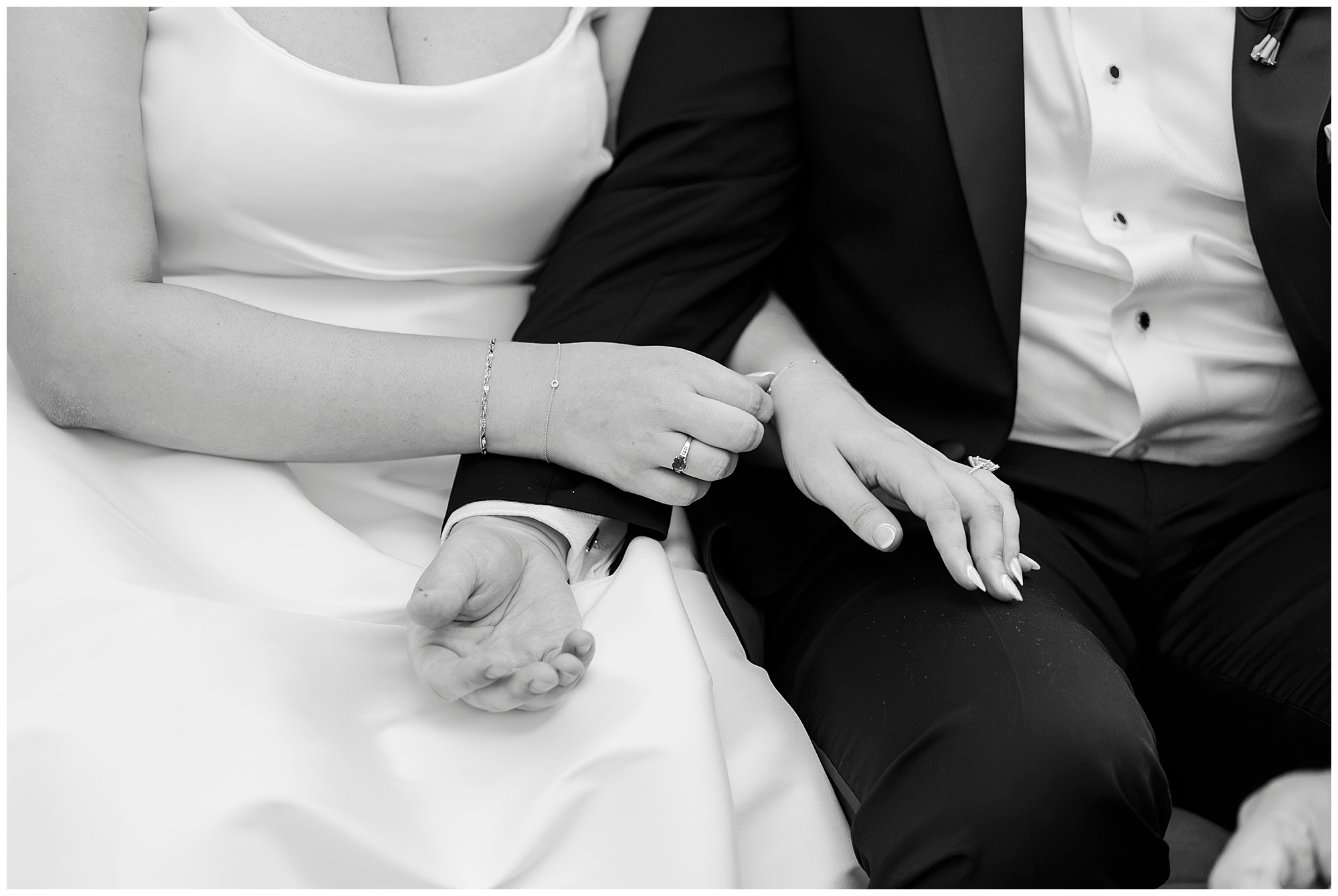 A bride and groom sit closely, holding hands. The bride, adorned in a white dress and bracelet, shares an intimate moment with the groom in his black suit and white shirt. Their focus is on each other’s hands, highlighting wedding rings—a timeless capture from a Felina wedding. Black and white photo.